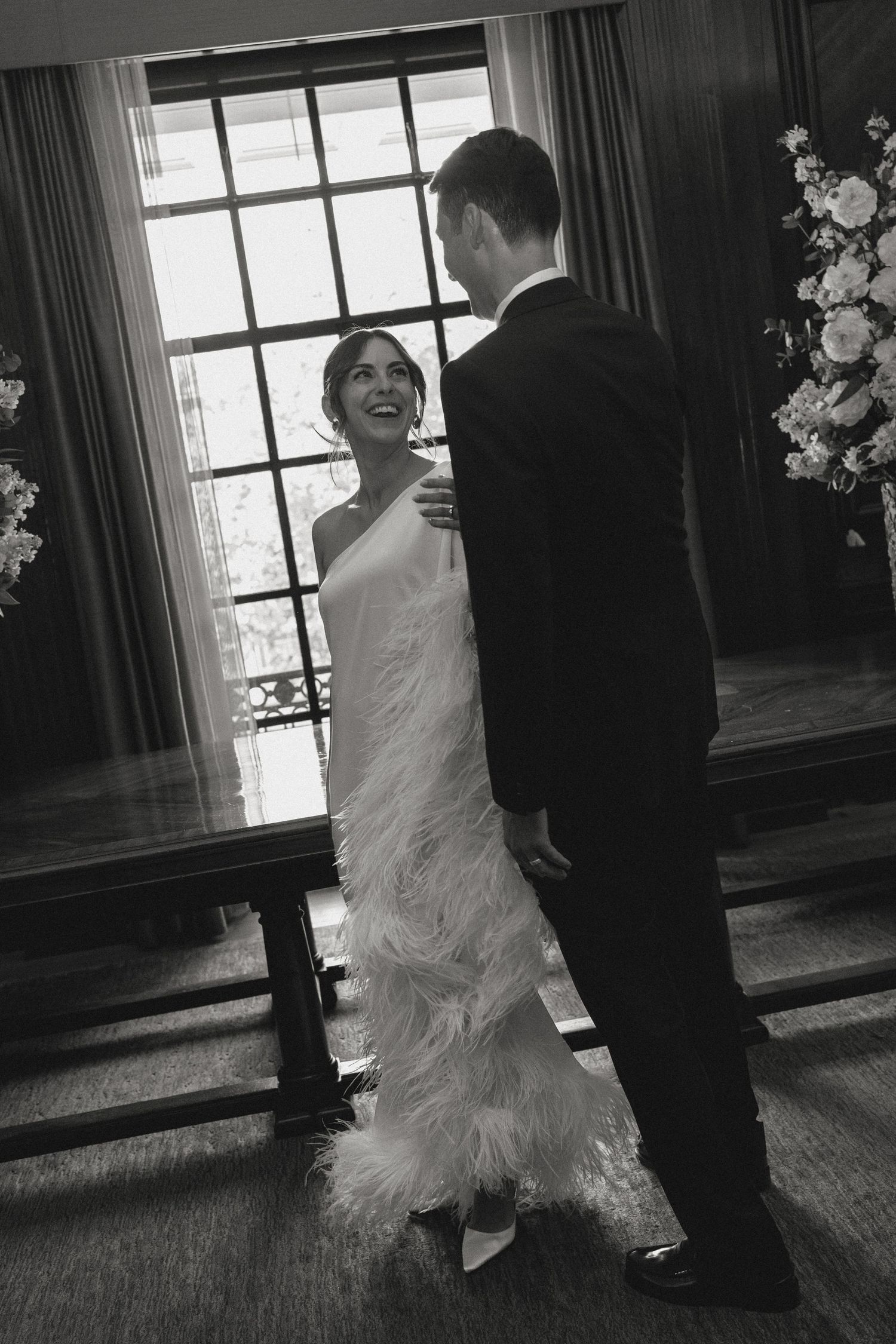 Black and white photo of couple dancing in elegant room with windows.