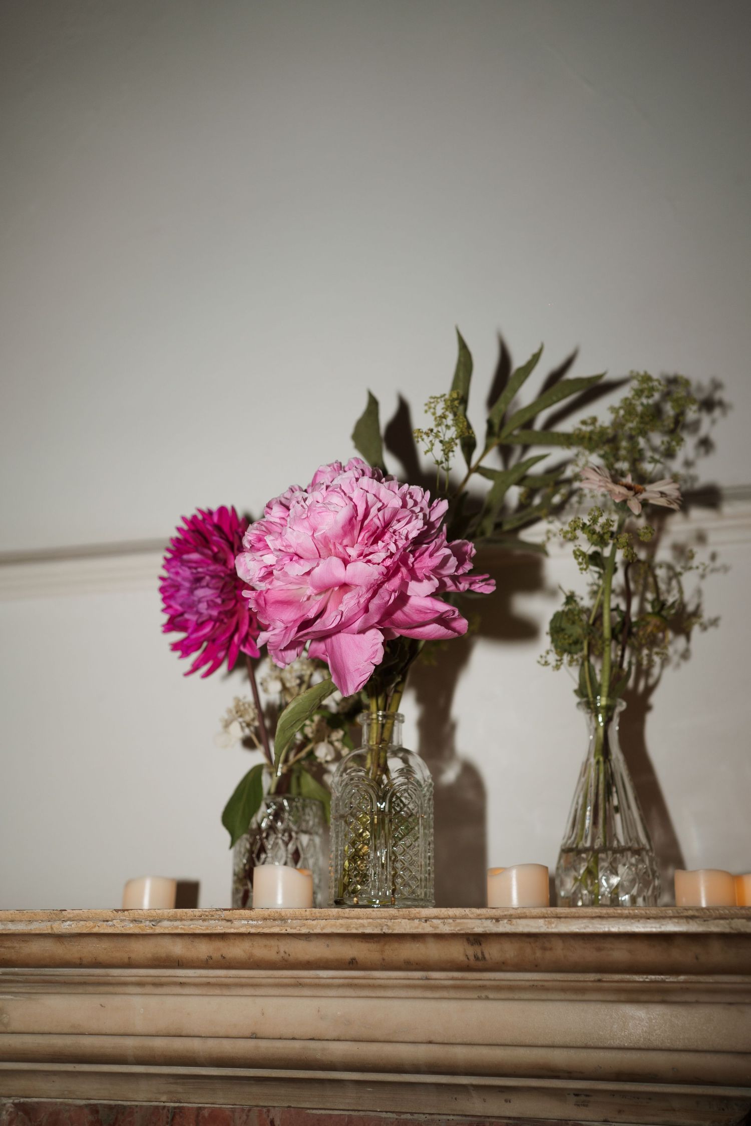 Pink and purple flowers arranged in glass vases with candles on a wooden mantel.