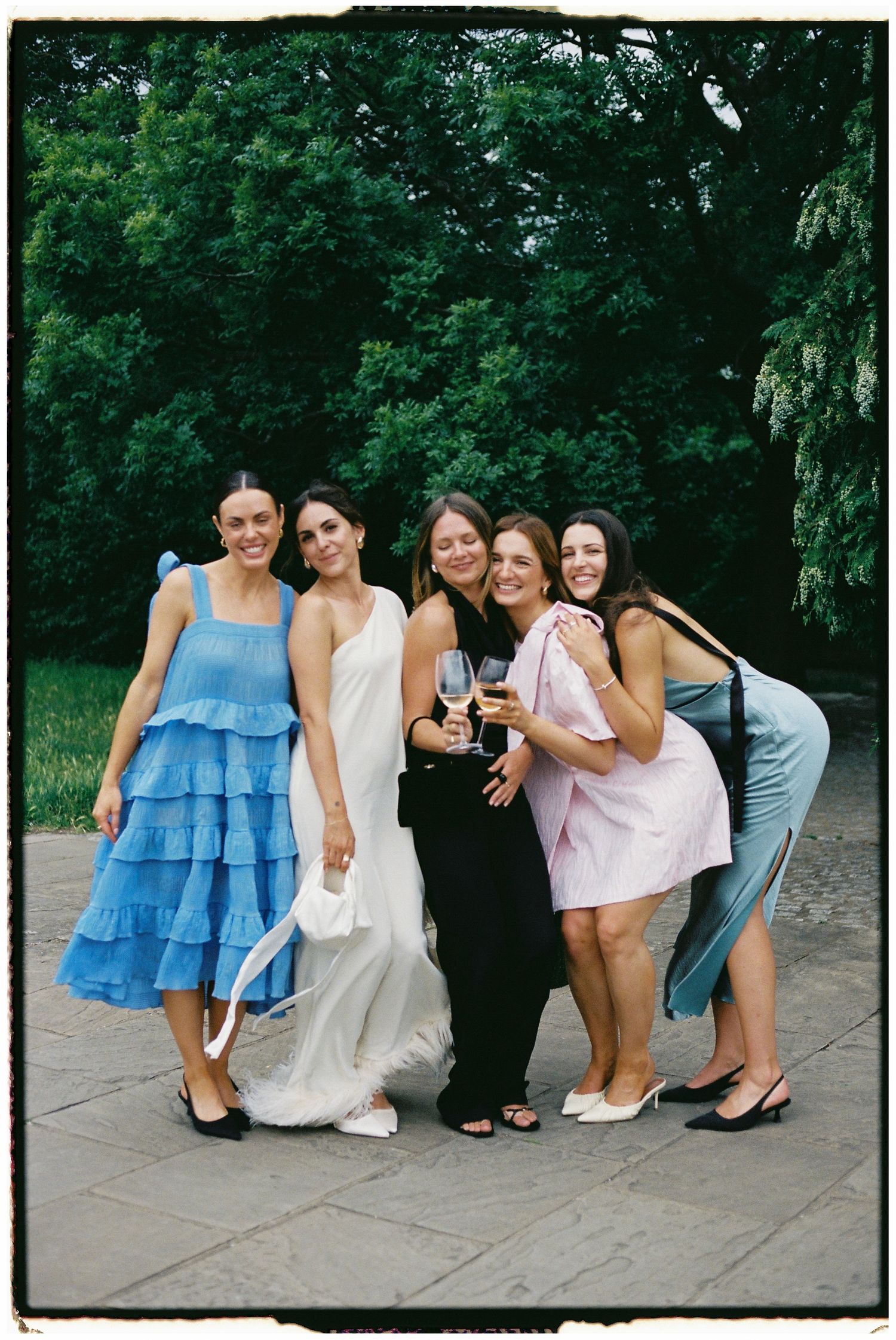 A group of friends in dresses pose together outdoors on a stone pathway.