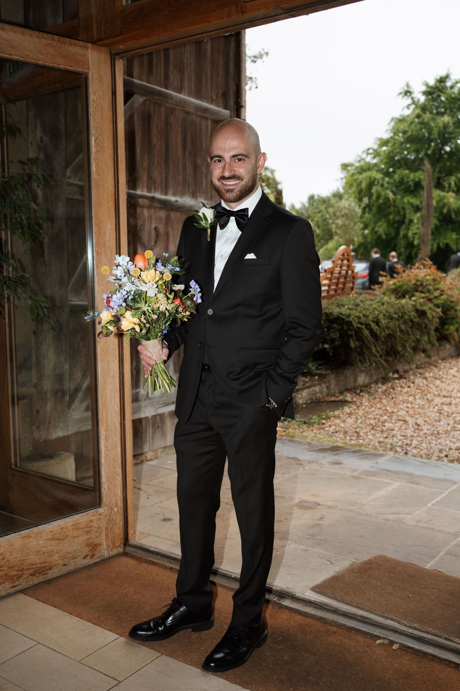 Groom in black tuxedo standing in rustic wooden doorway.