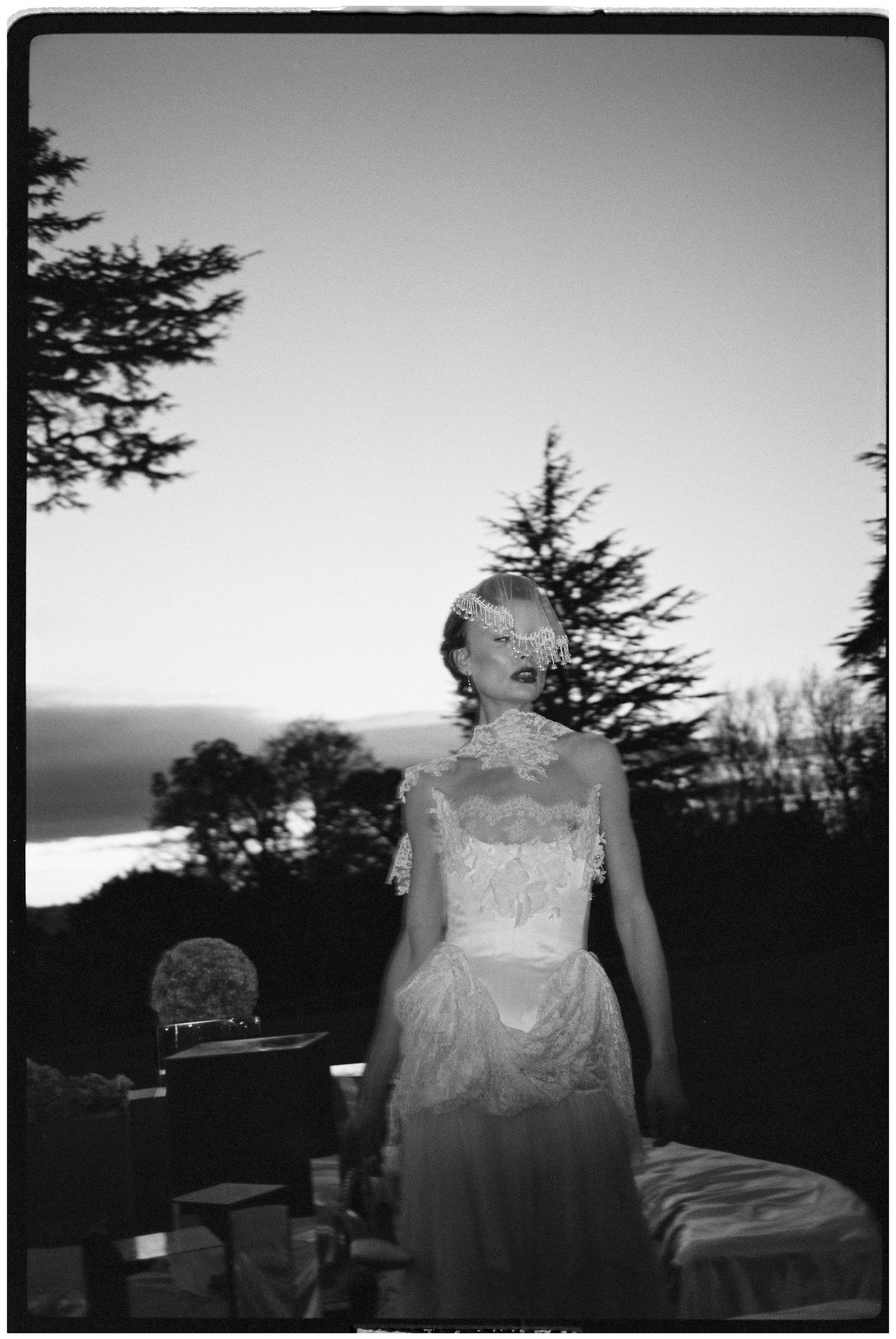 Ethereal black and white portrait of bride in lace dress against sunset and pine trees.