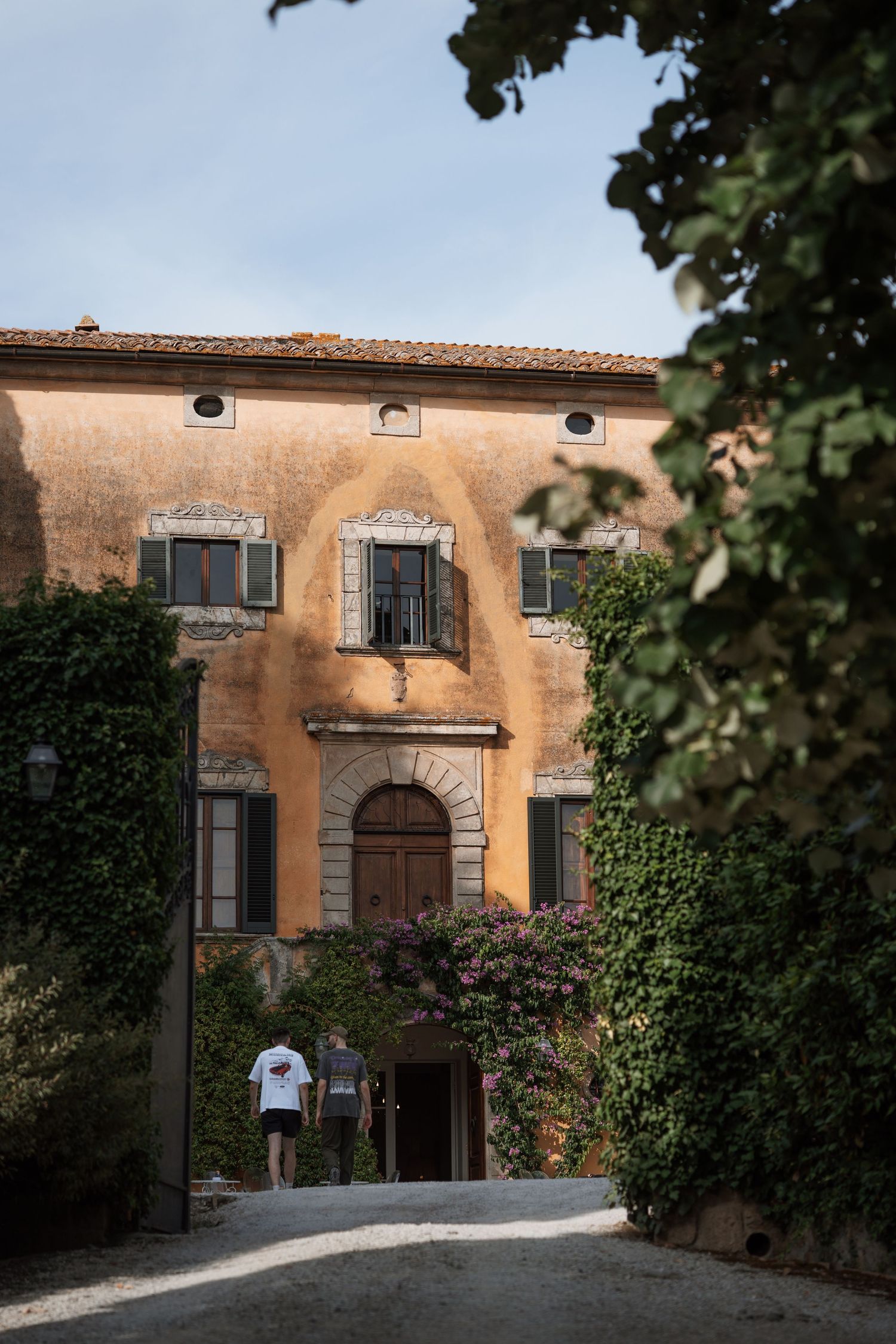 Two people walking toward an ivy-covered Italian villa with ornate arched doorway and weathered ochre facade.