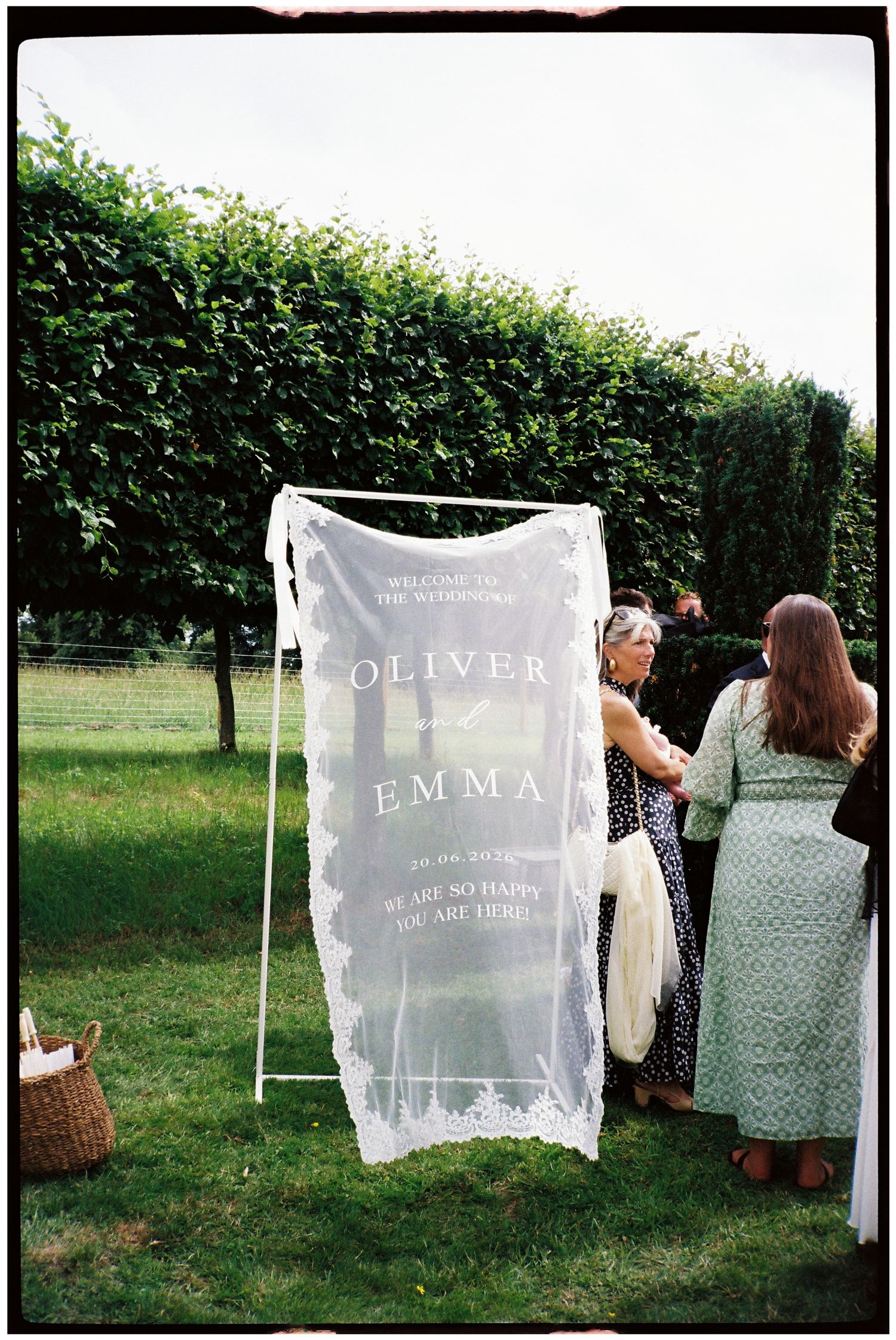 Outdoor wedding welcome sign on sheer lace fabric displaying Oliver and Emma names on green hedgerow backdrop.