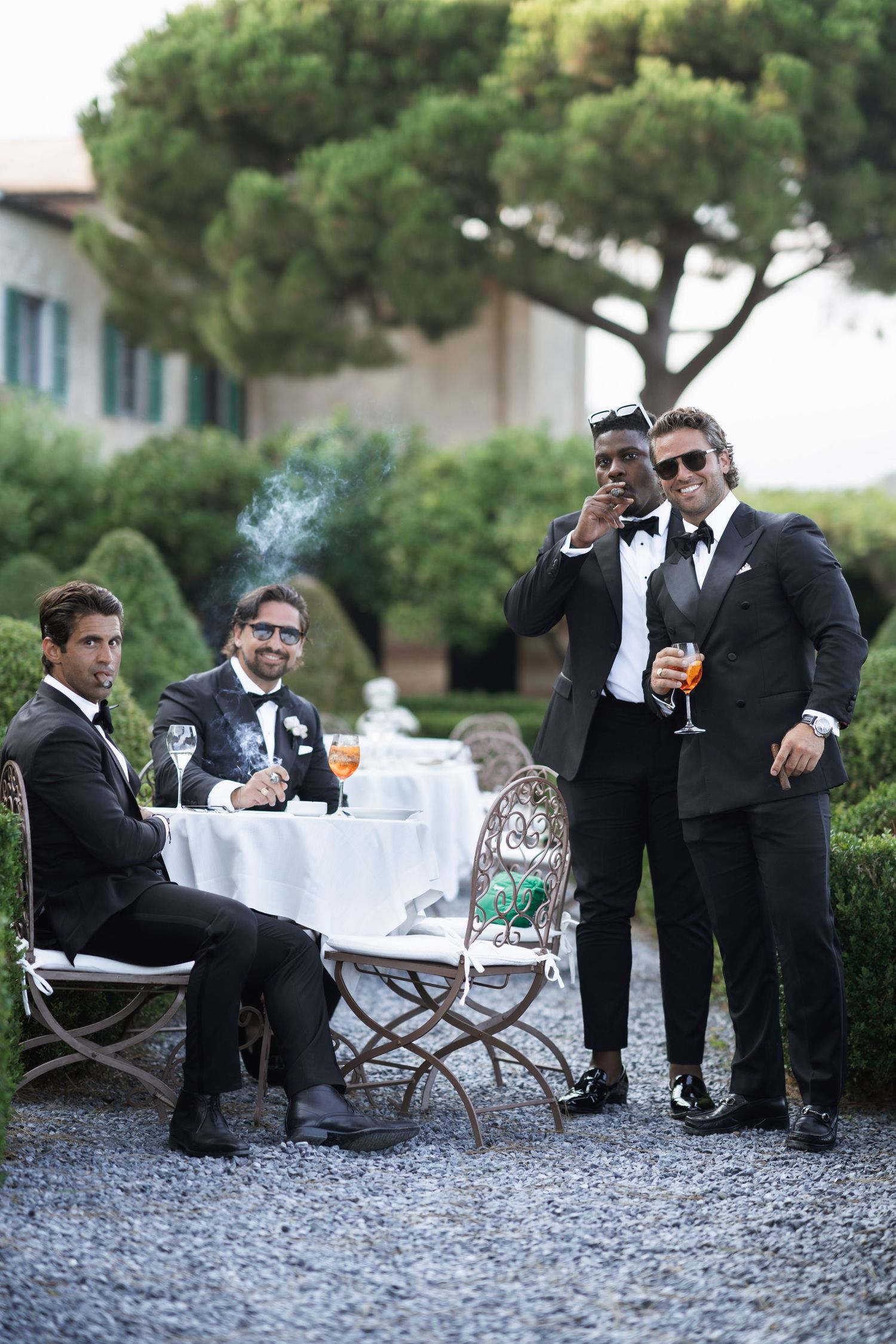 Groomsmen in black tuxedos relaxing on stone patio with drinks.