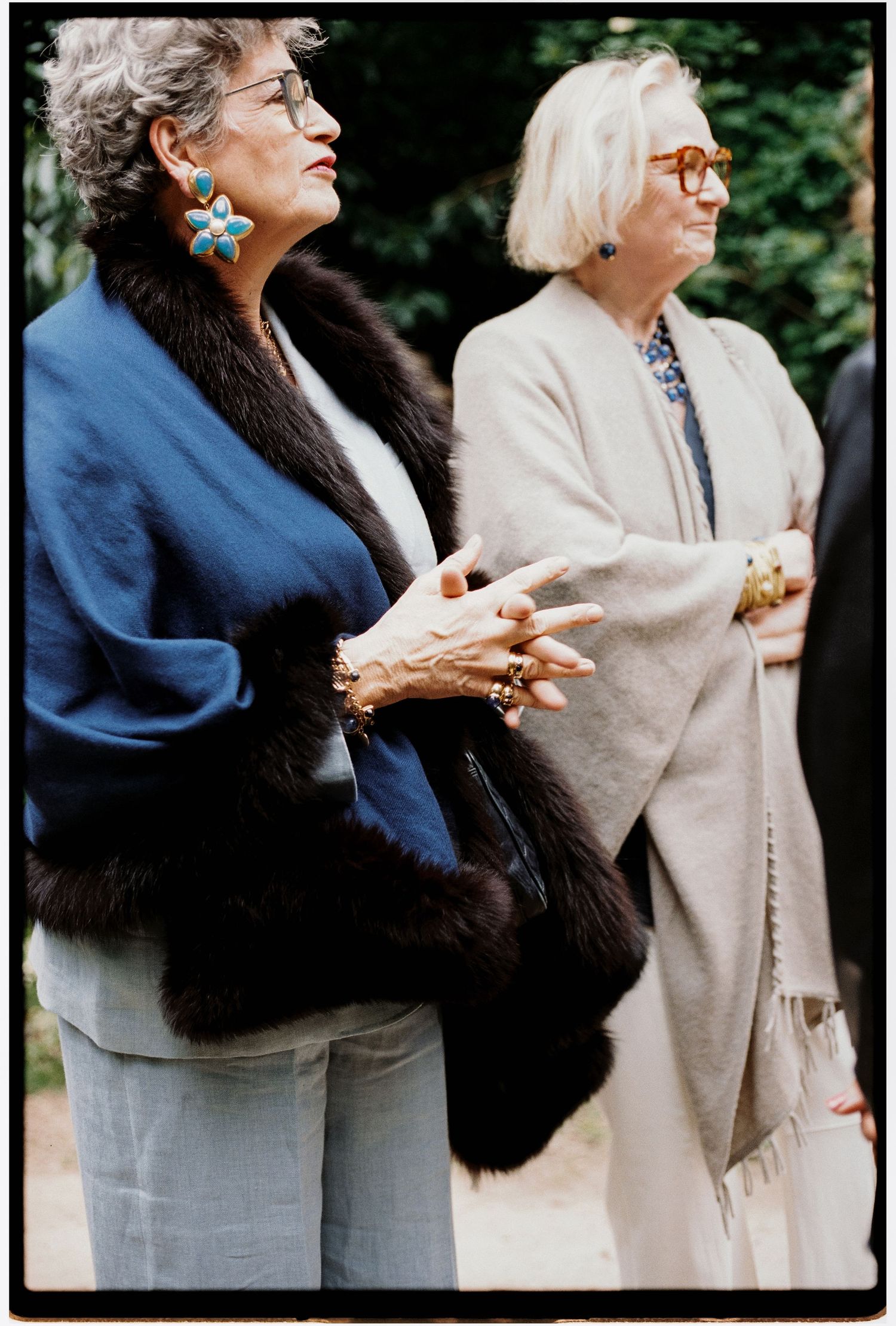 Two elegant older women in formal attire, one in blue with fur stole, one in beige wrap, wearing glasses outdoors.