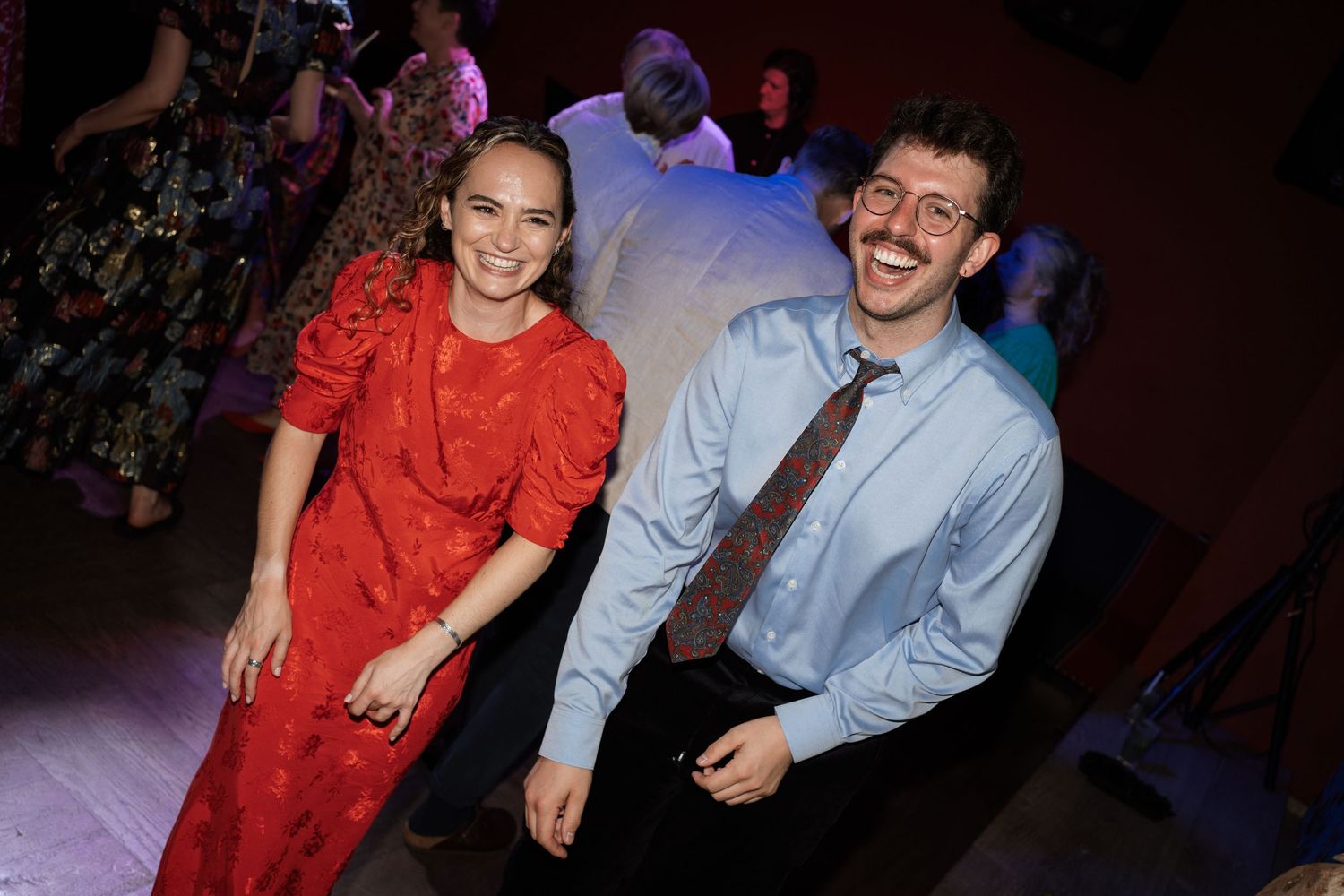 A couple dancing and laughing together at a party, with the woman wearing a red dress and man in blue shirt.