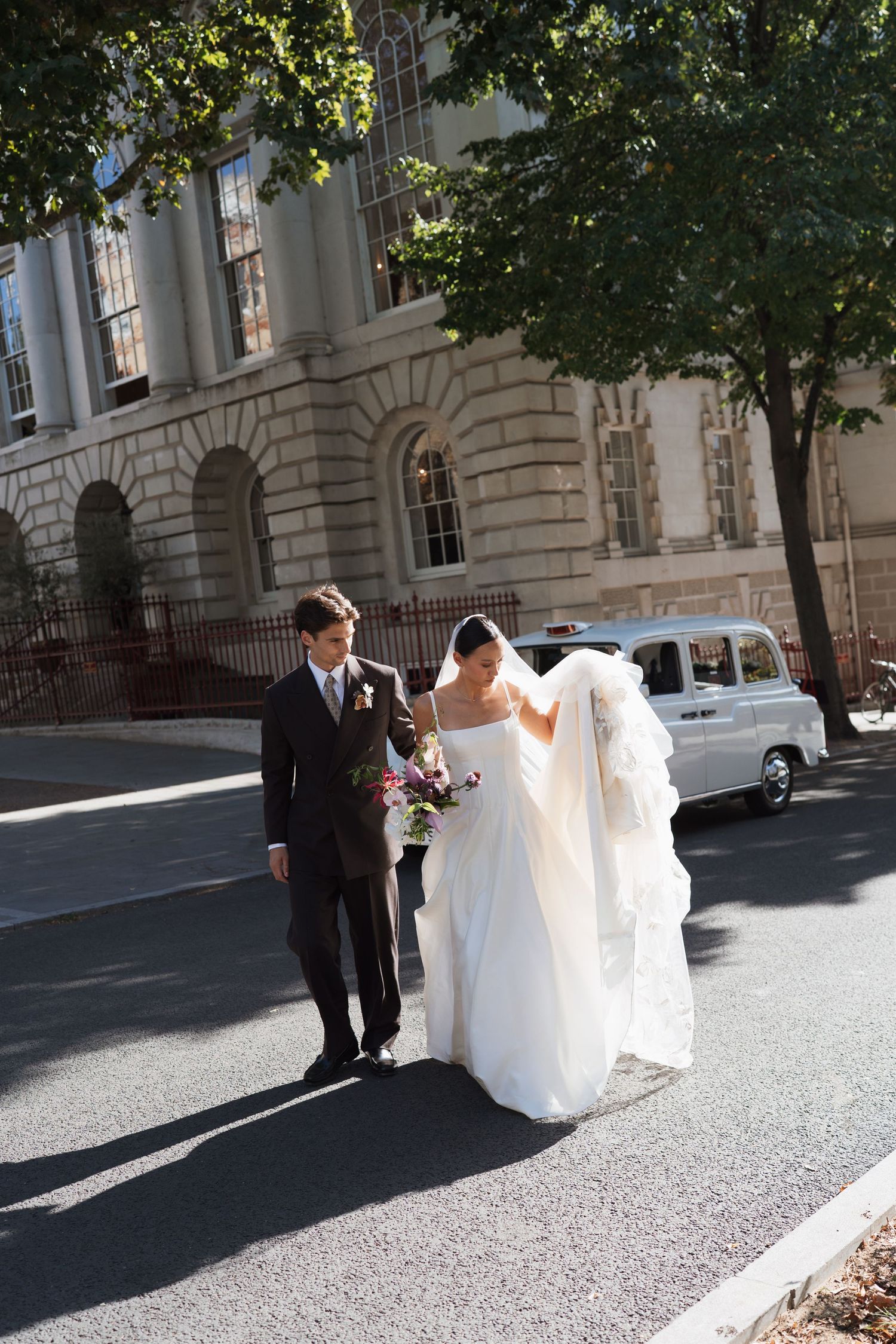 A bride in a white gown and veil walks beside a groom in a dark suit on a sunlit street near a historic stone building.