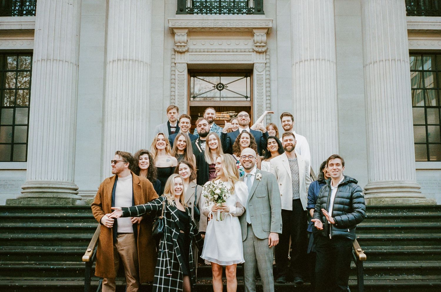 Large wedding party poses for group photo on historic building steps with classical columns.