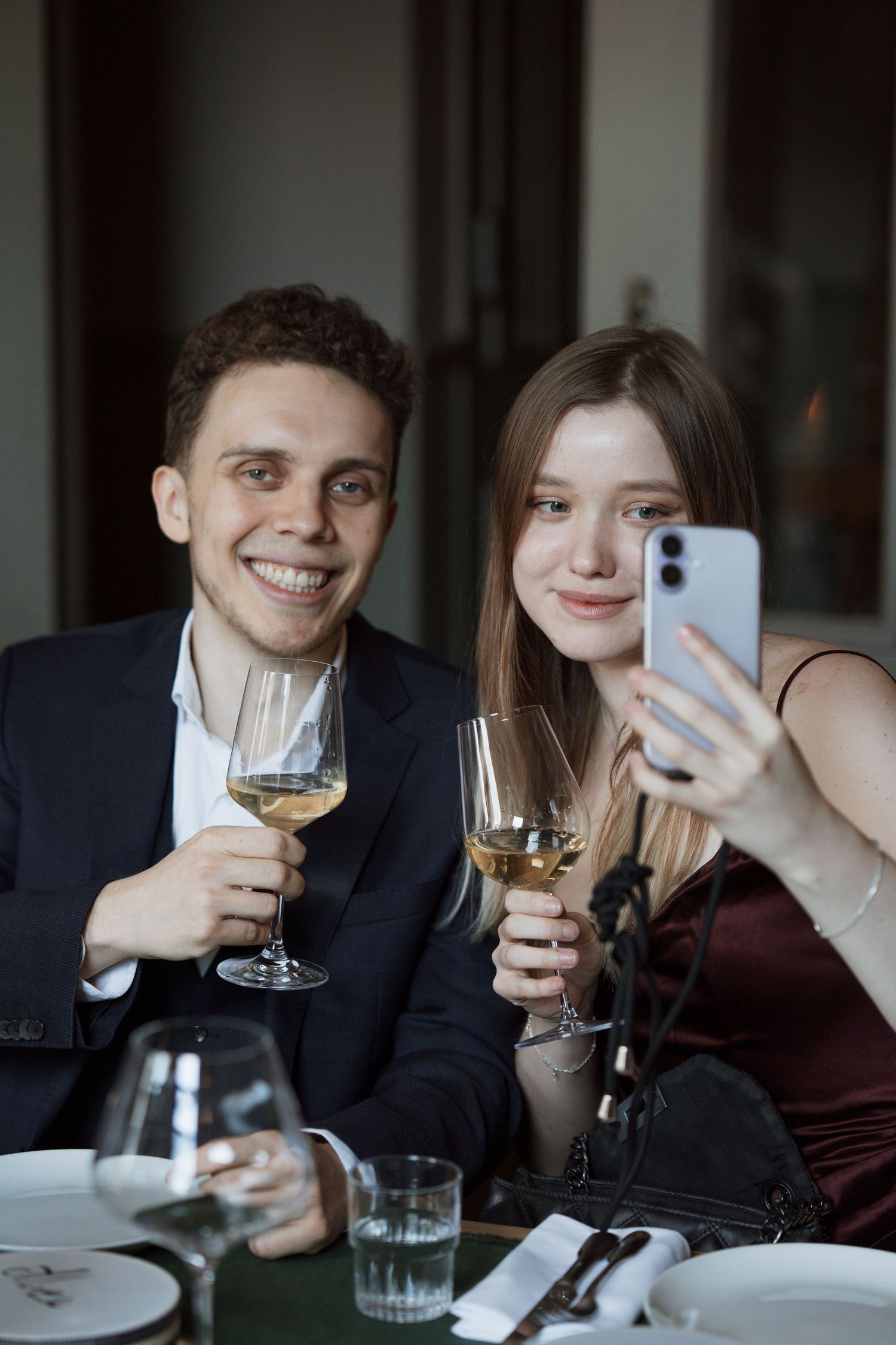 Two people taking selfies while enjoying wine at a restaurant table.