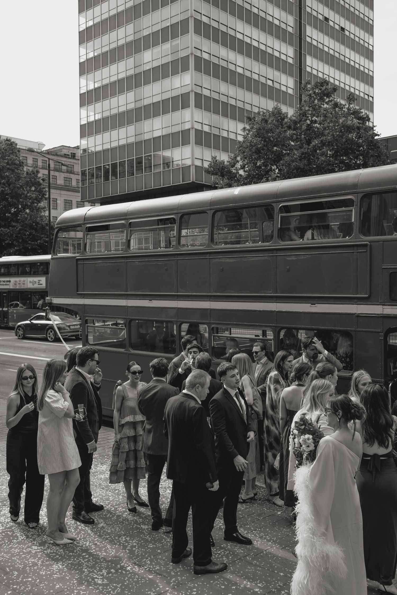 Large group of wedding guests gathered near vintage buses outside an office building.
