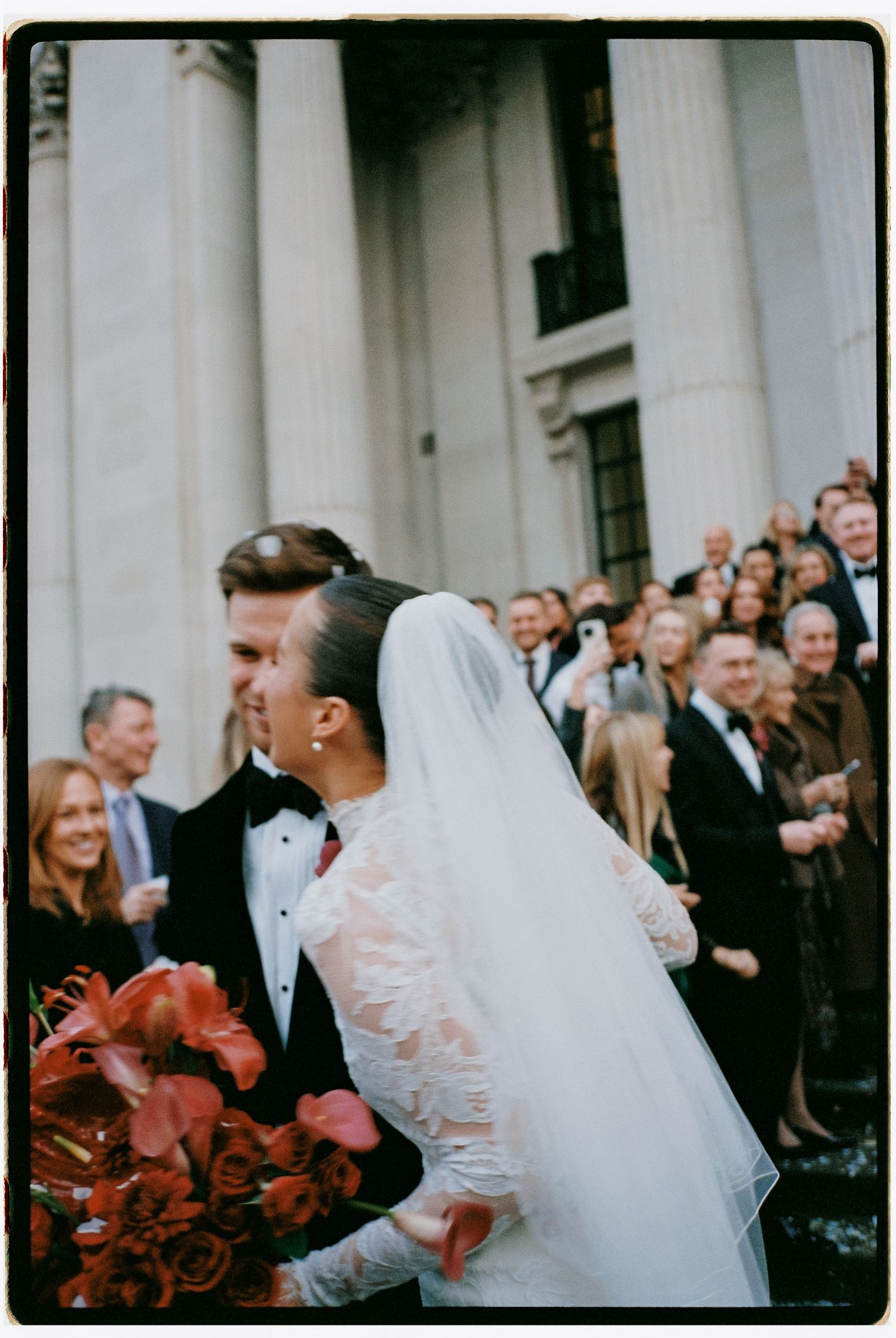 Newlyweds exiting church ceremony with classical columns and crowd of guests in background.