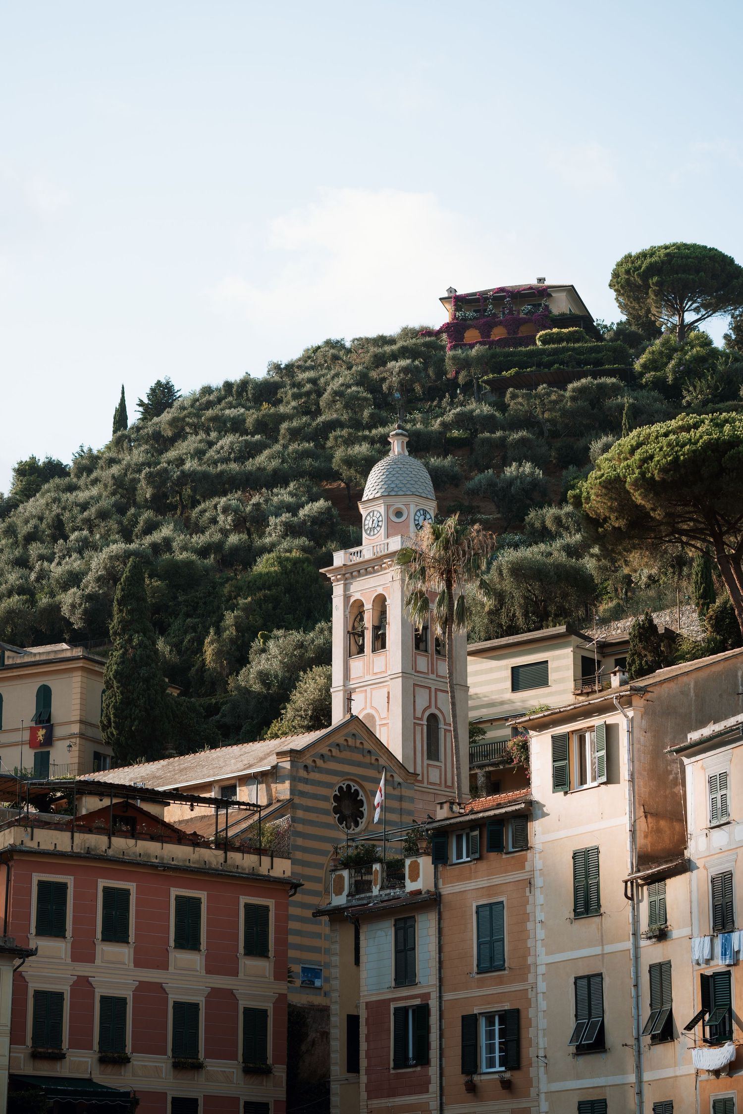 Pastel colored Italian coastal buildings with bell tower nestled on hillside among trees.