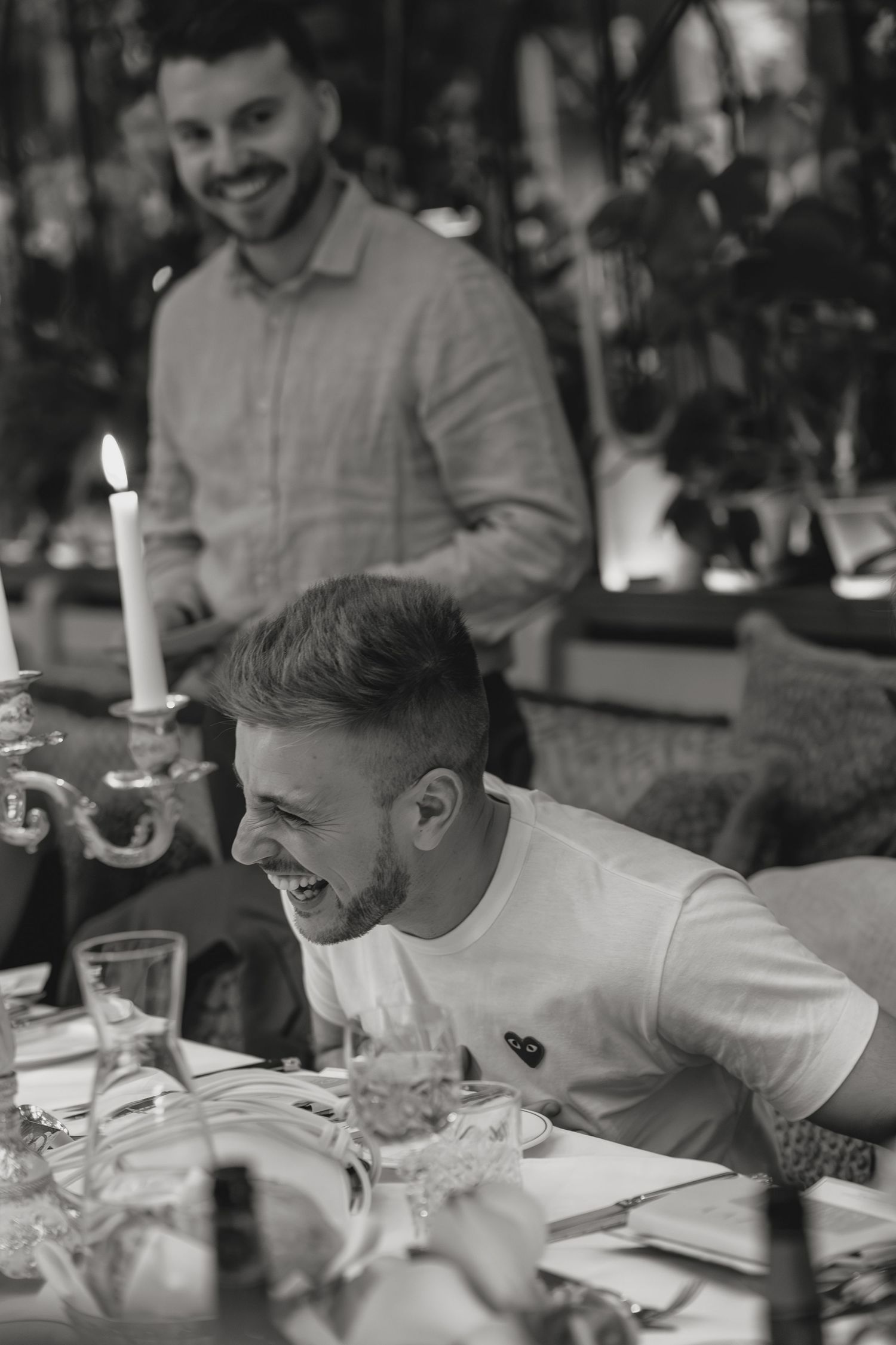 Black and white photo of two smiling men at an elegant candlelit dinner table in a restaurant.