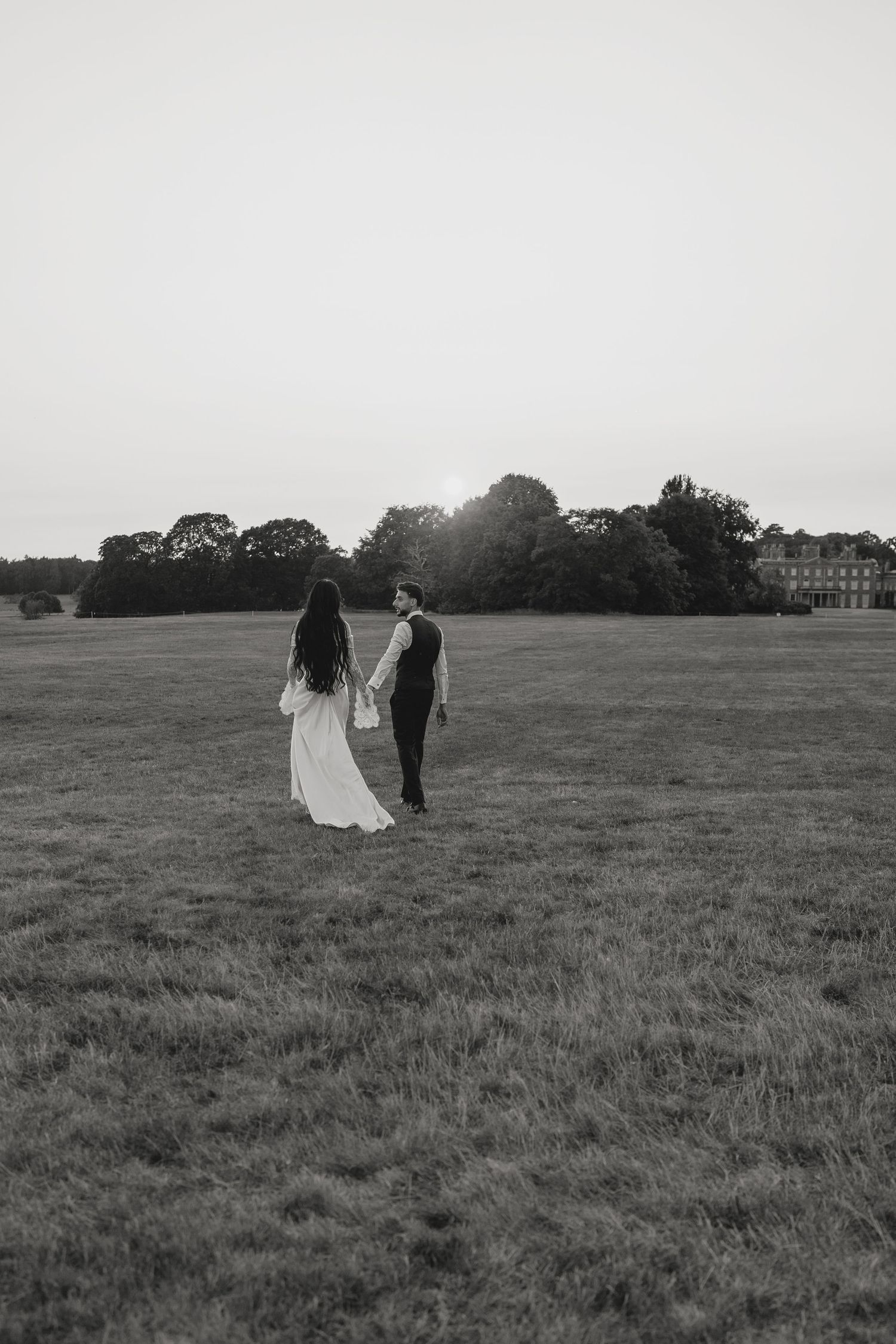 A couple walks hand in hand across a vast open field in moody black and white.