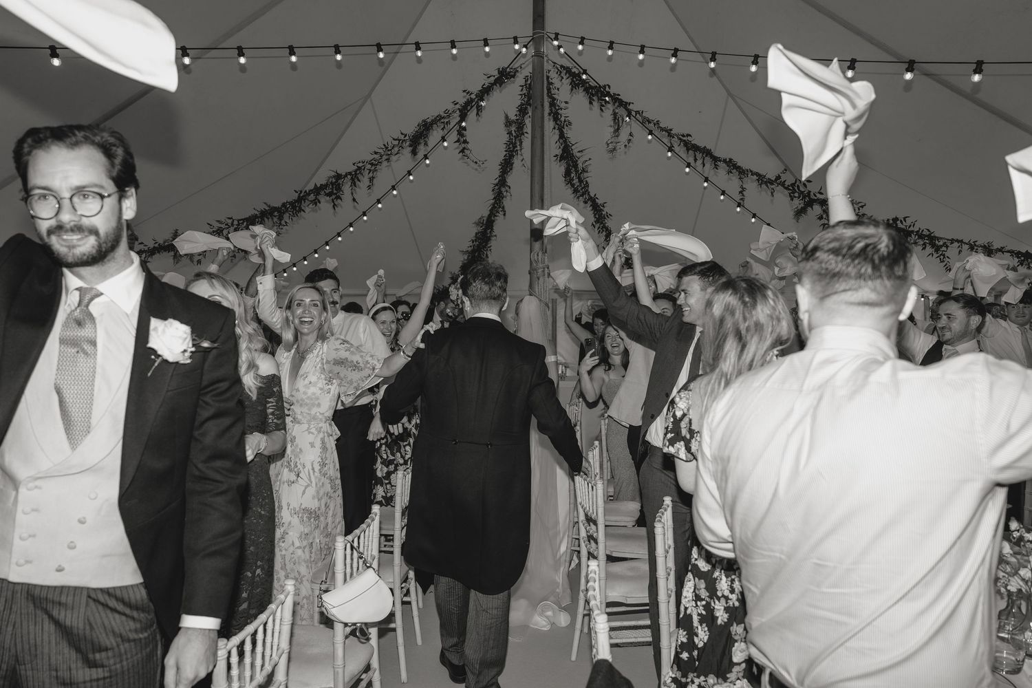 Black and white wedding photo of guests celebrating in a marquee, waving napkins as couple walks through aisle.