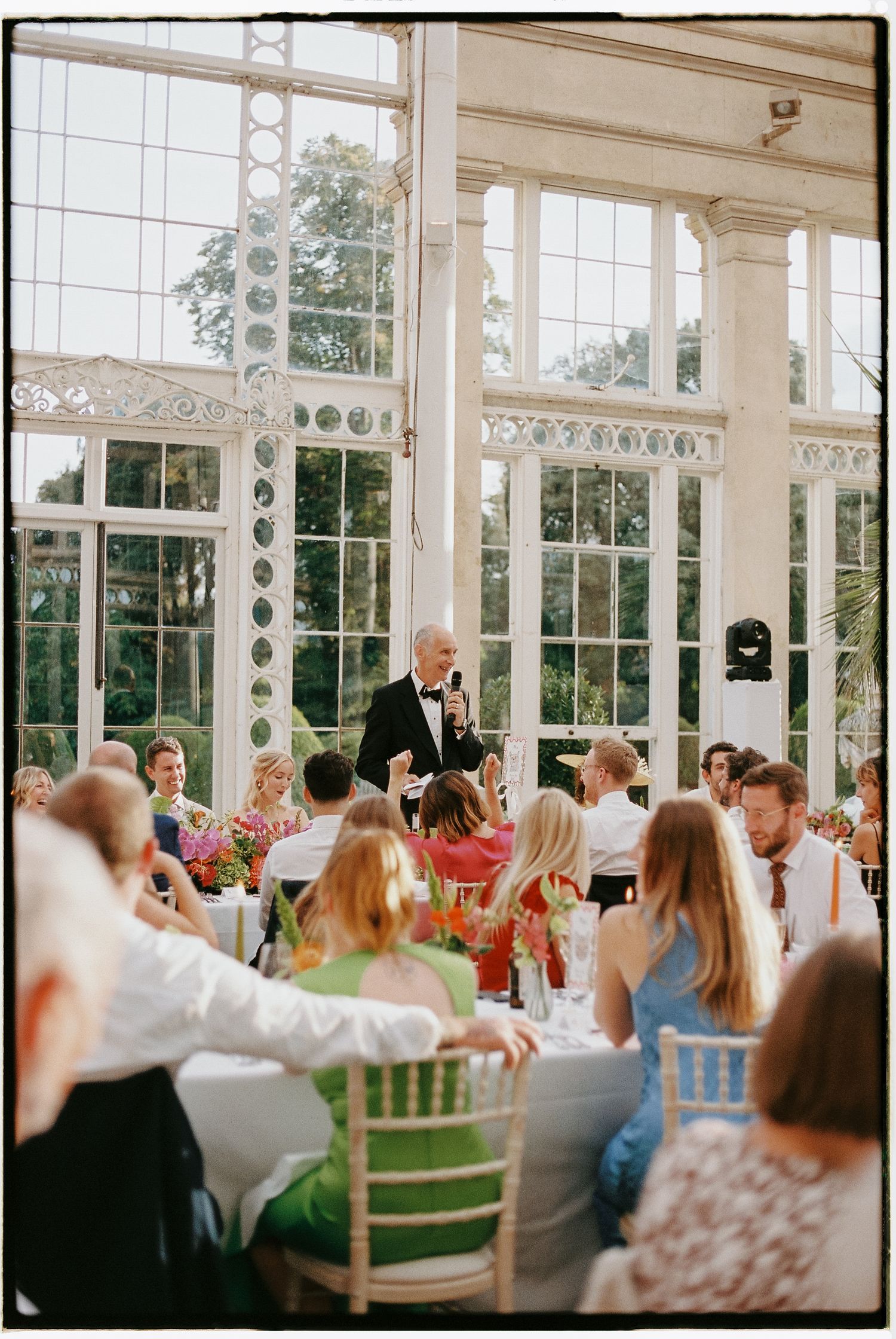 Man in tuxedo giving speech at elegant greenhouse wedding reception with guests seated at decorated tables.