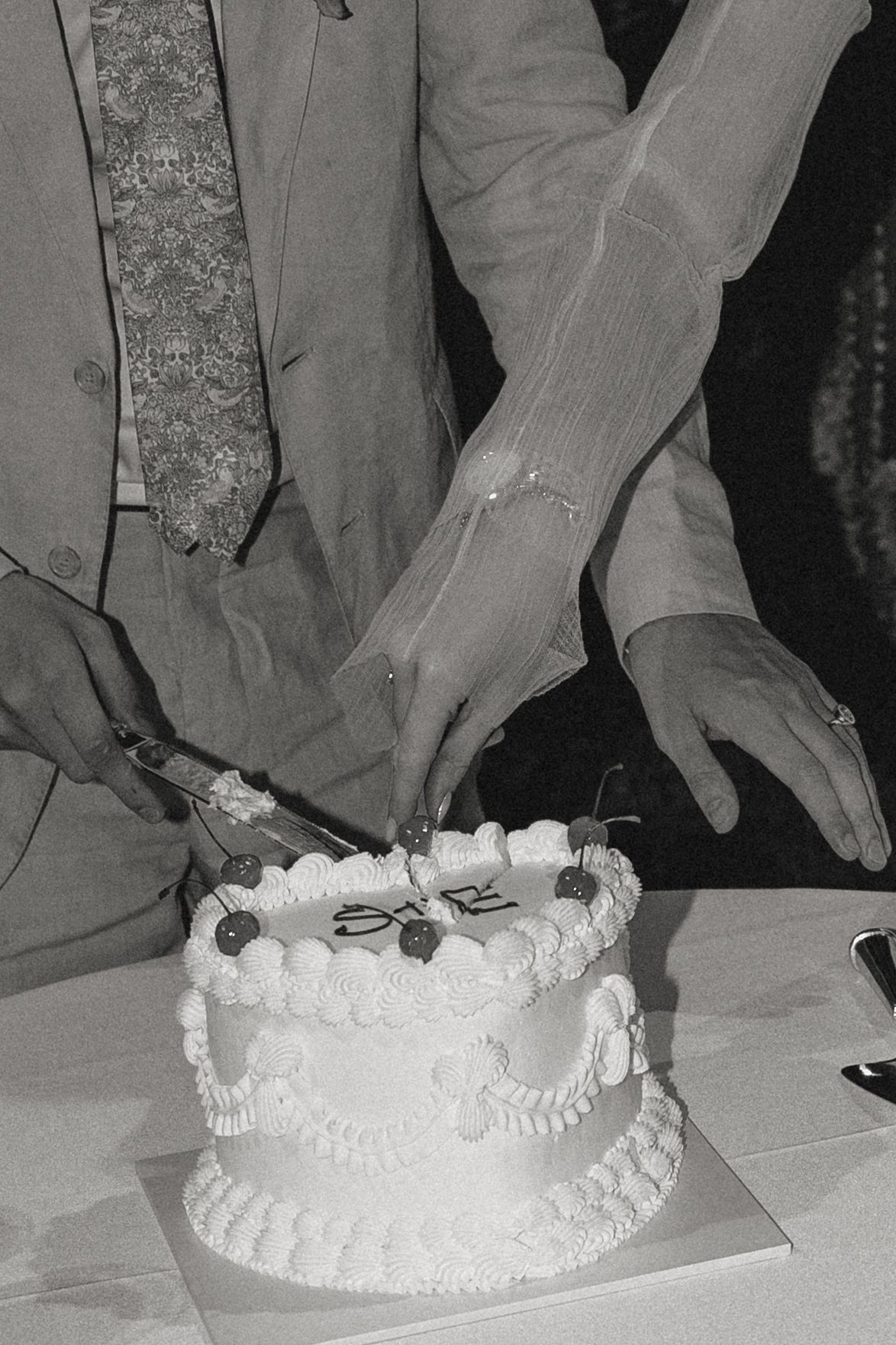 Black and white photo of hands cutting into an ornate white wedding cake.