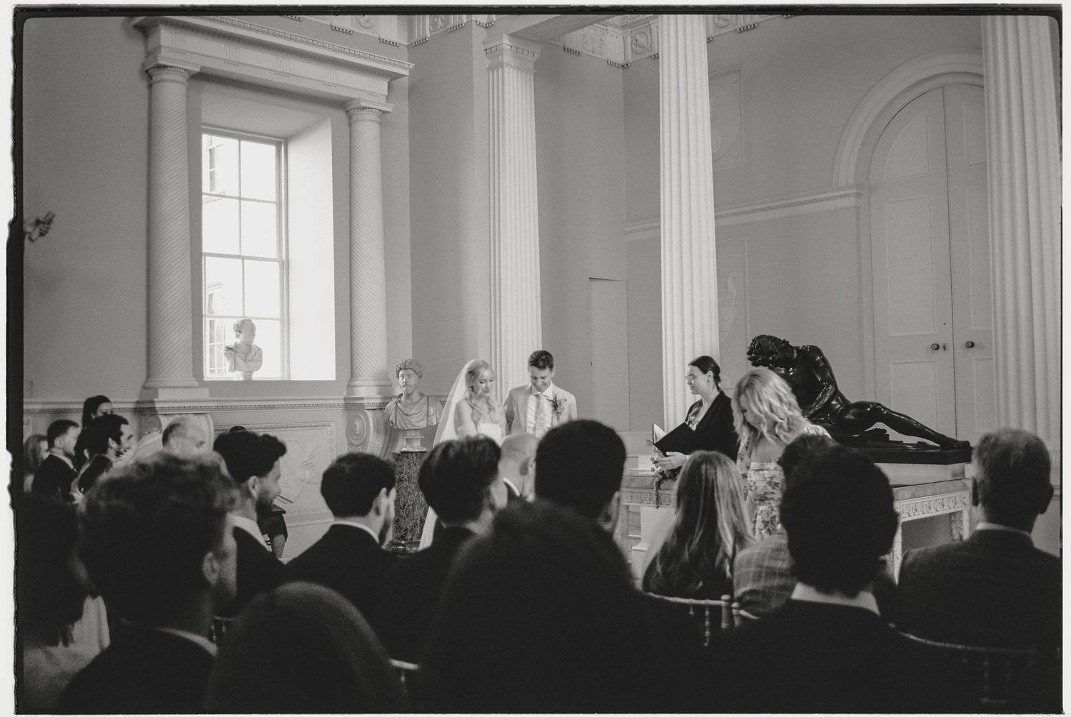 Black and white photo of a formal gathering in an elegant columned hall with an audience watching a ceremony.