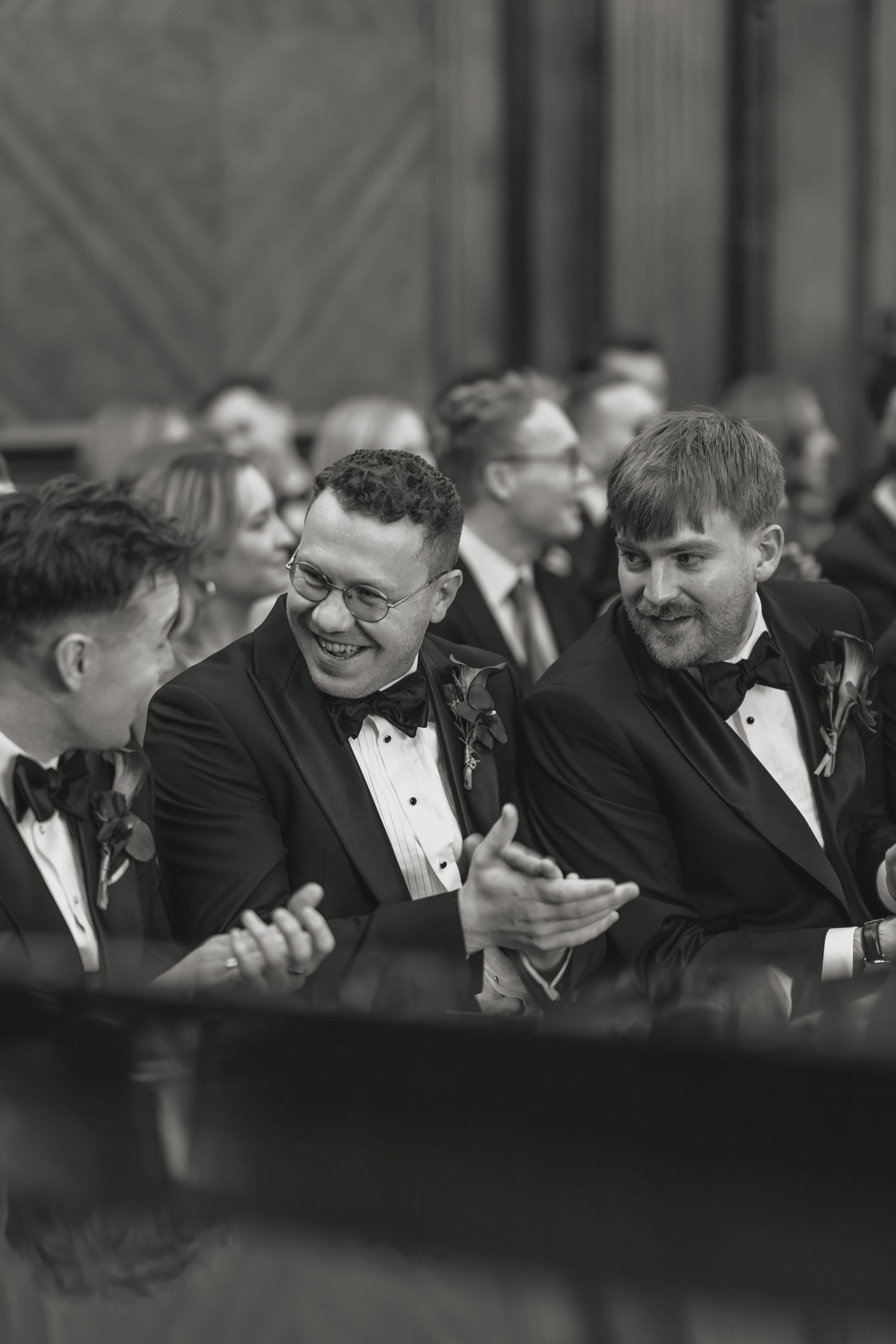 Groomsmen share laughs and check phones during candid black and white moment.