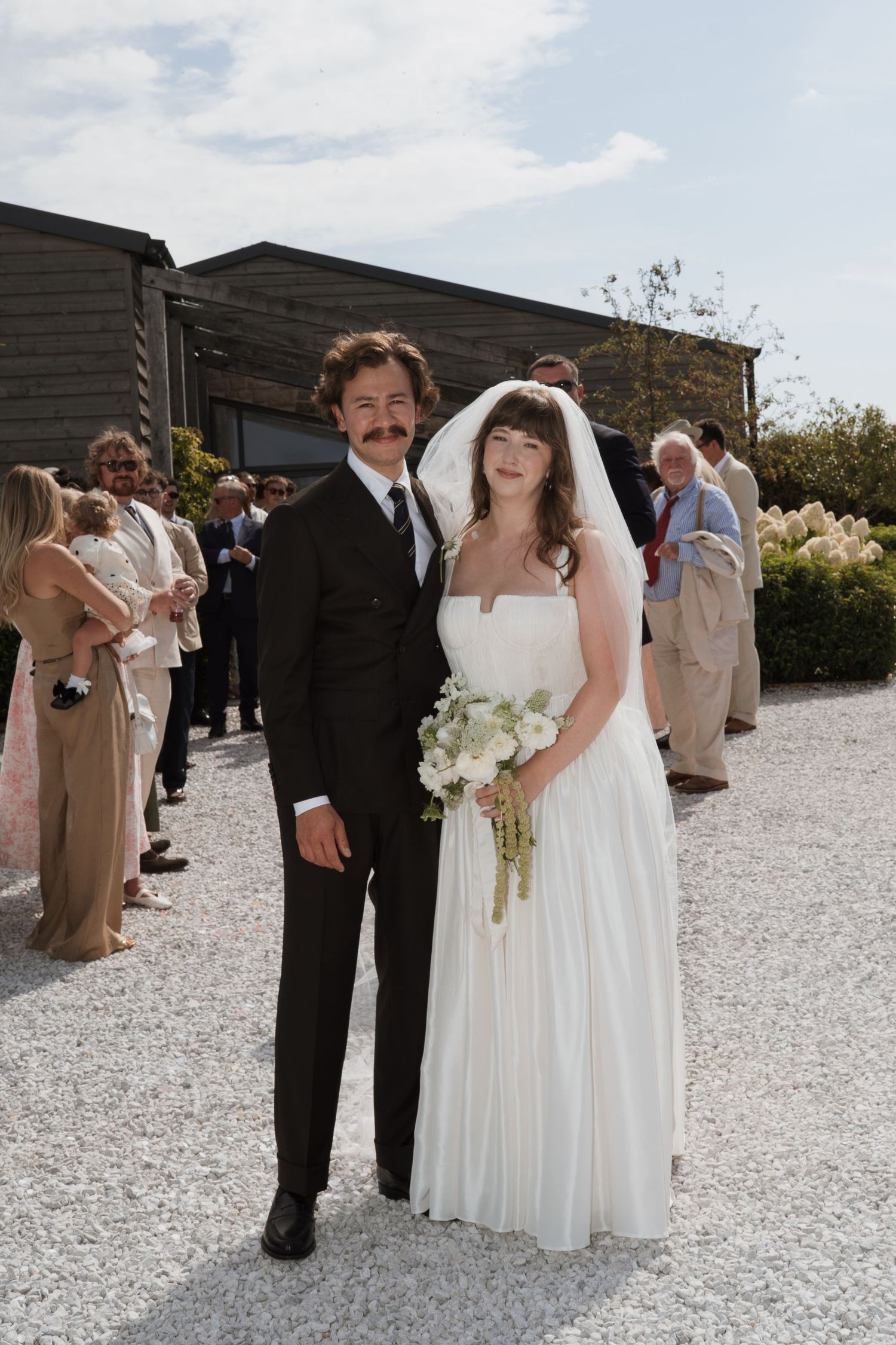 Newlyweds pose on gravel path with wedding party in beige and black attire.