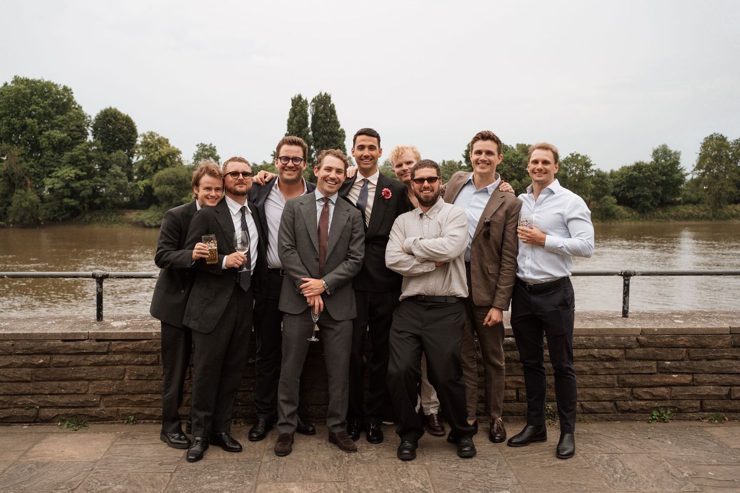 Wedding party members sit together on a stone wall near a river.