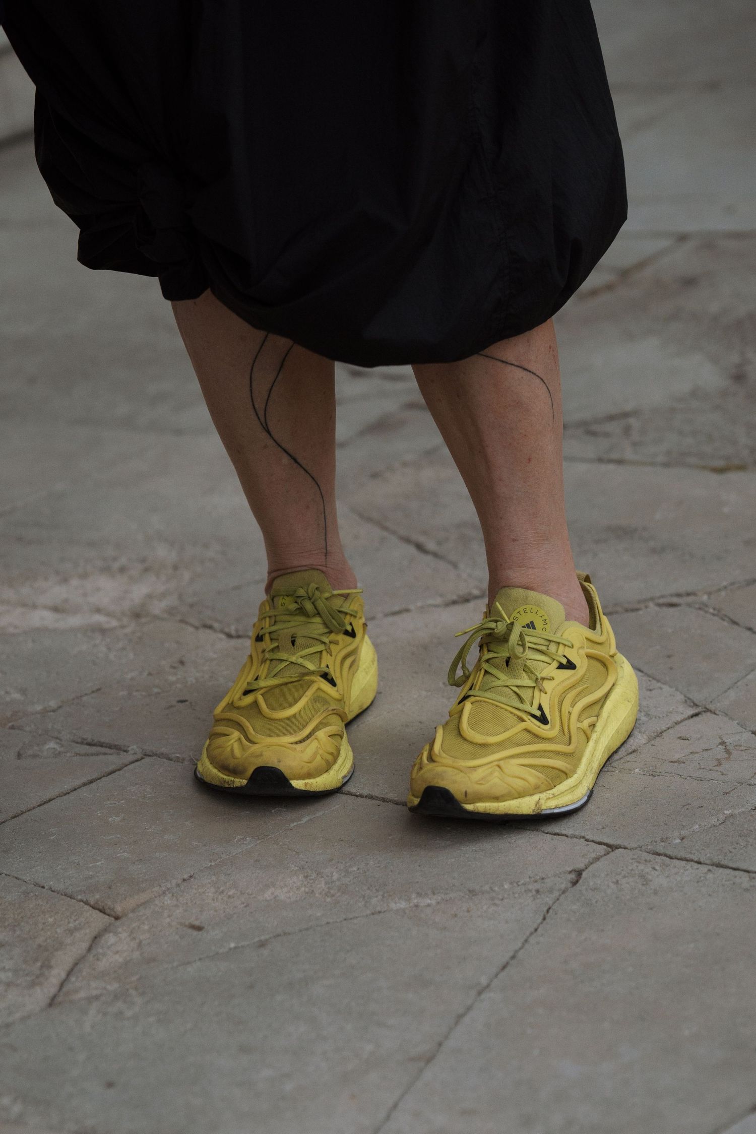 Bright yellow athletic shoes worn with formal black attire on stone flooring.