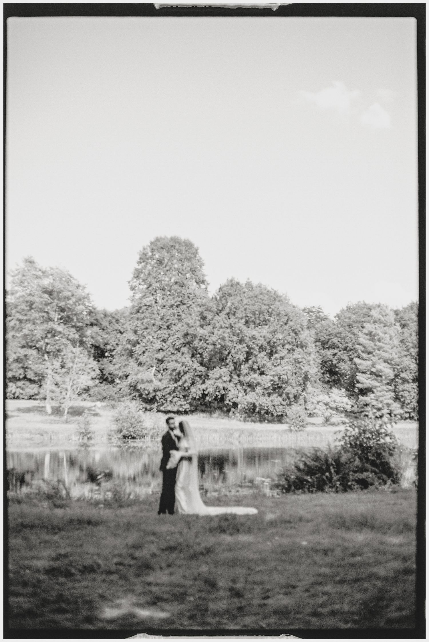 Black and white wedding photo of couple by lake with trees in background.