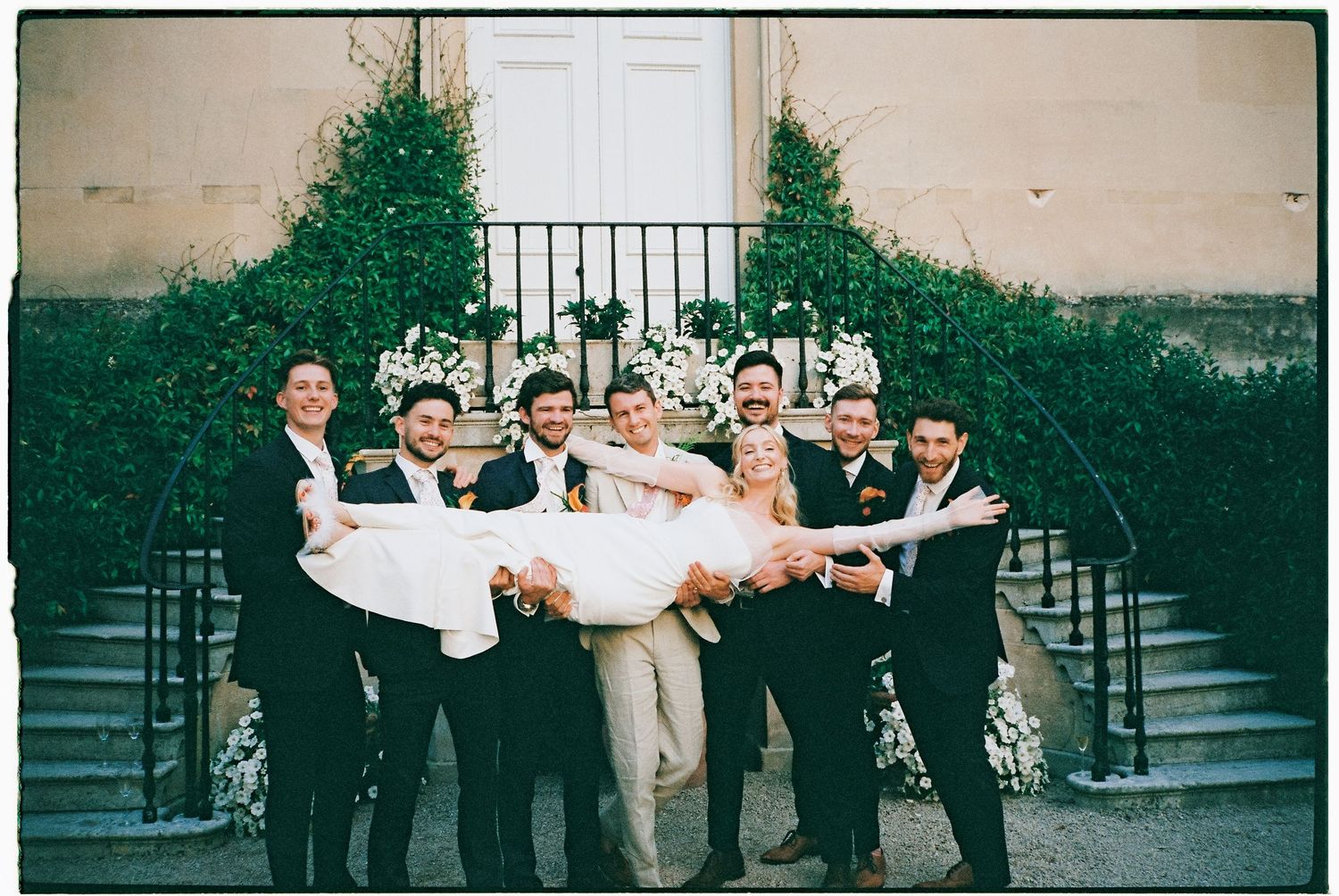 A group of formally dressed people pose together against a stone wall with greenery.