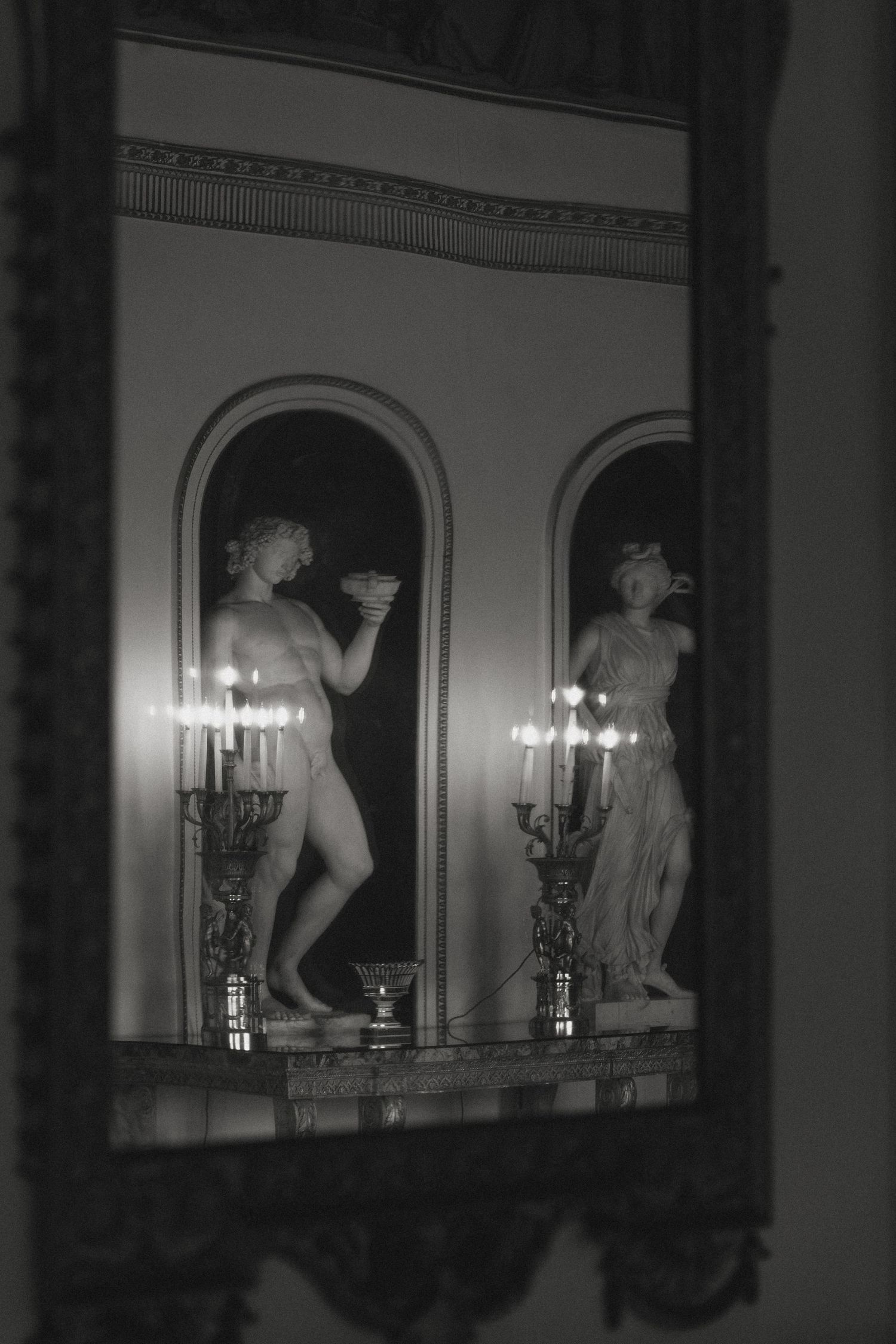 Classical statues and candelabras line an ornate mirrored wall in a dimly lit room.