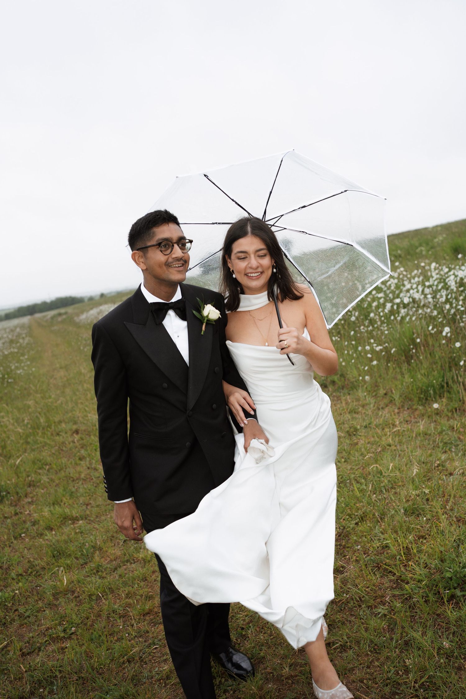 A couple poses in a grassy field with a clear umbrella on their wedding day.