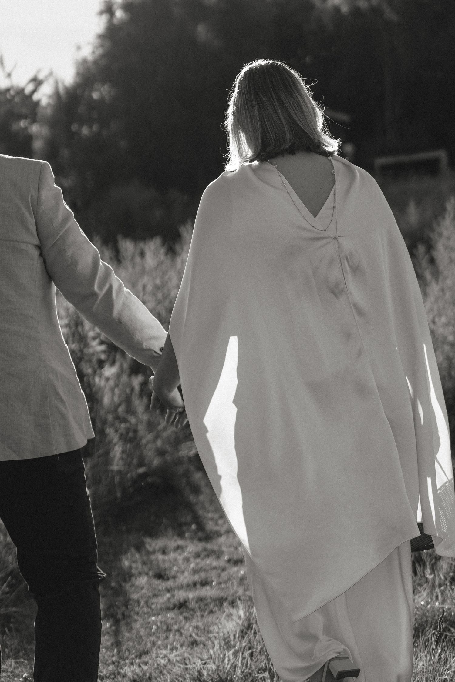 Black and white photo of a couple holding hands walking outdoors, woman in flowing white dress, man in light suit.