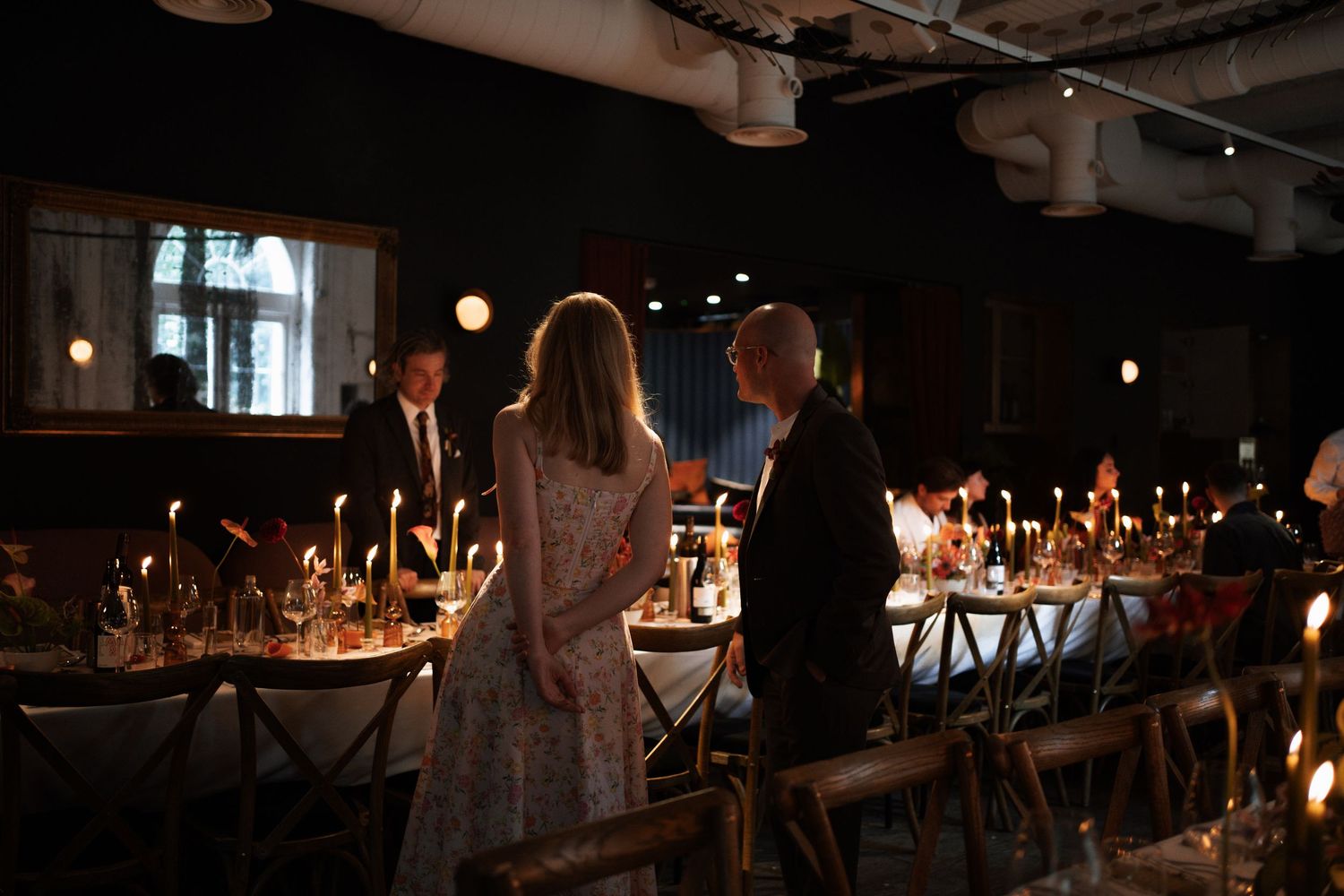 Elegant candlelit dinner event with guests mingling at long decorated tables in a dimly lit venue.