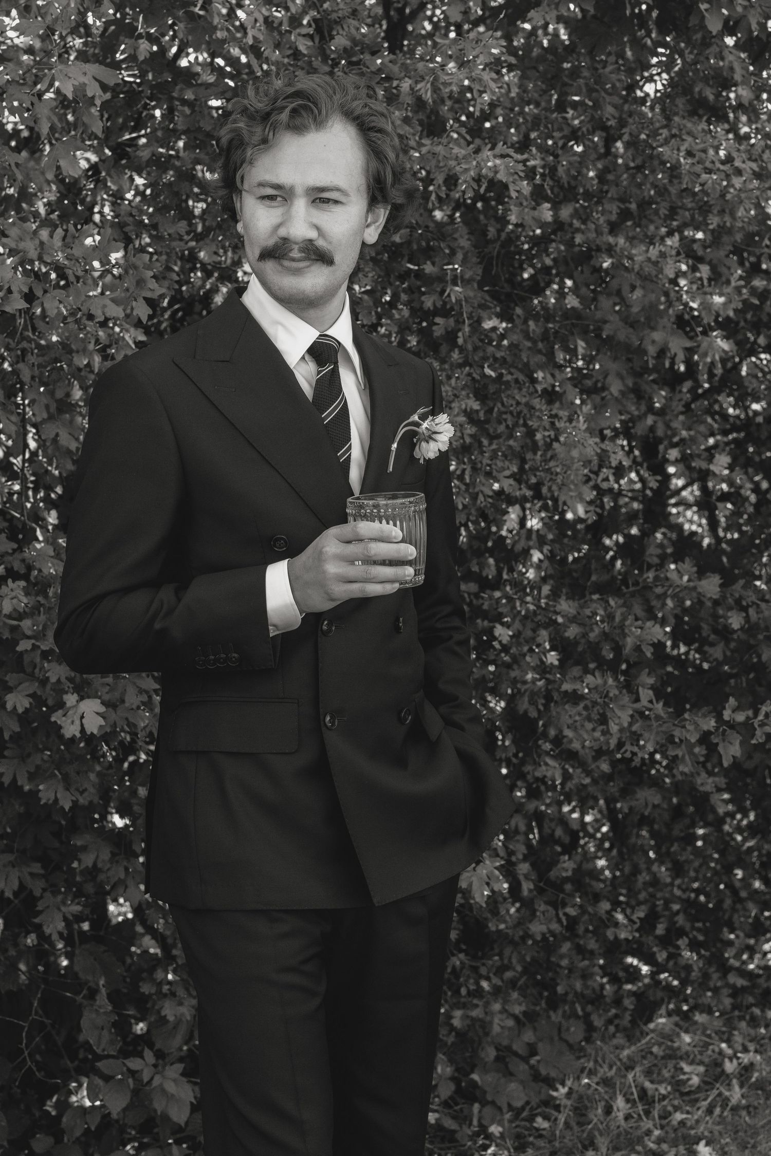 A person in a black suit poses with a drink against a leafy backdrop.