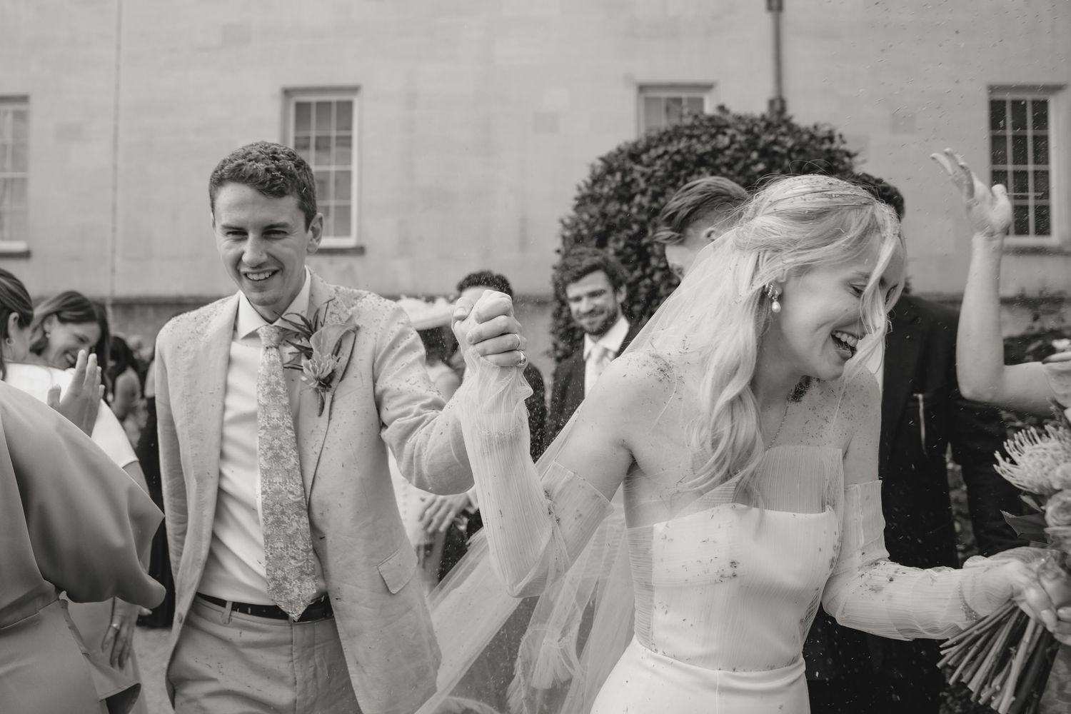 Wedding party members celebrate joyfully in front of a historic stone building.