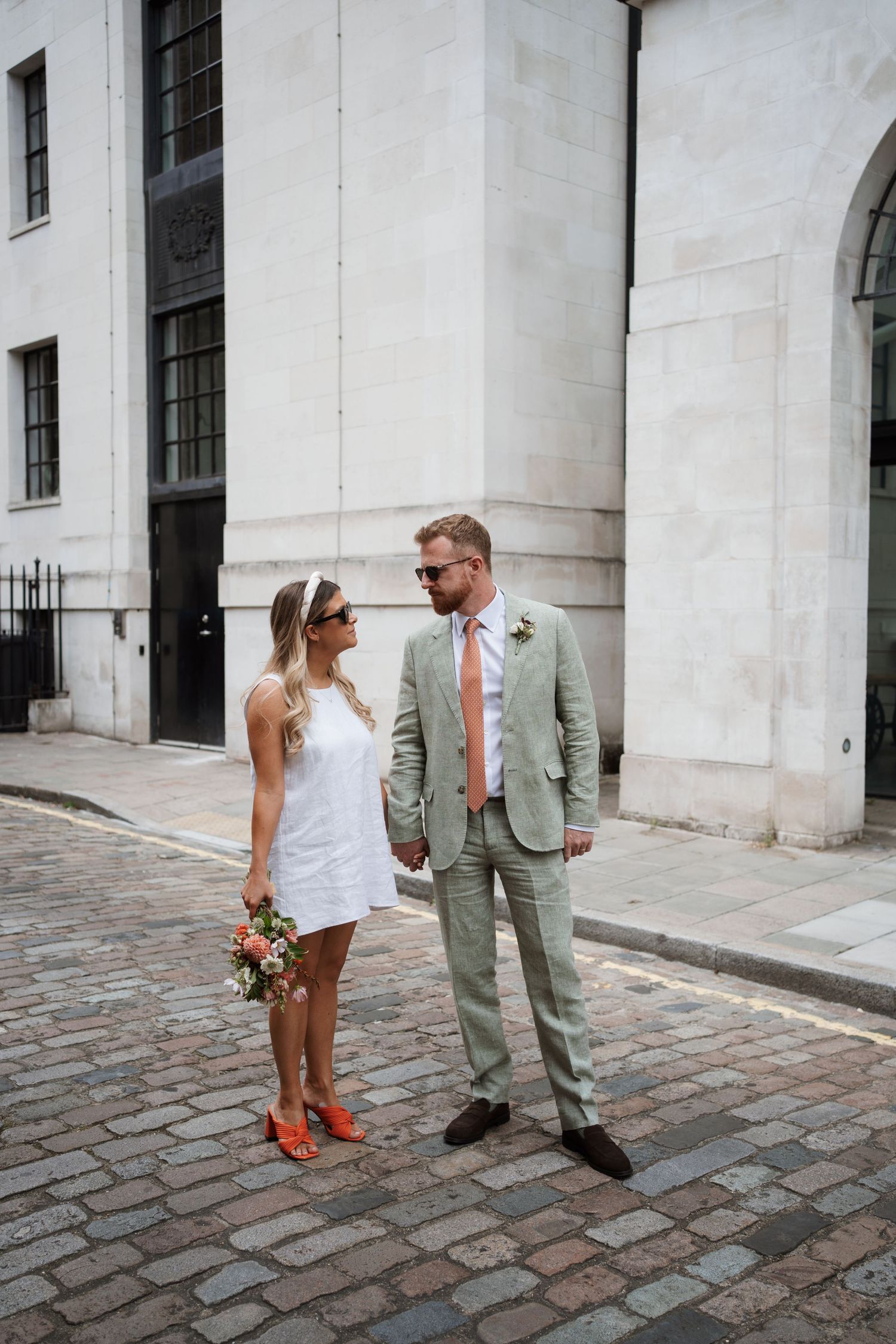 Couple in wedding attire walks on cobblestone street near white stone building.