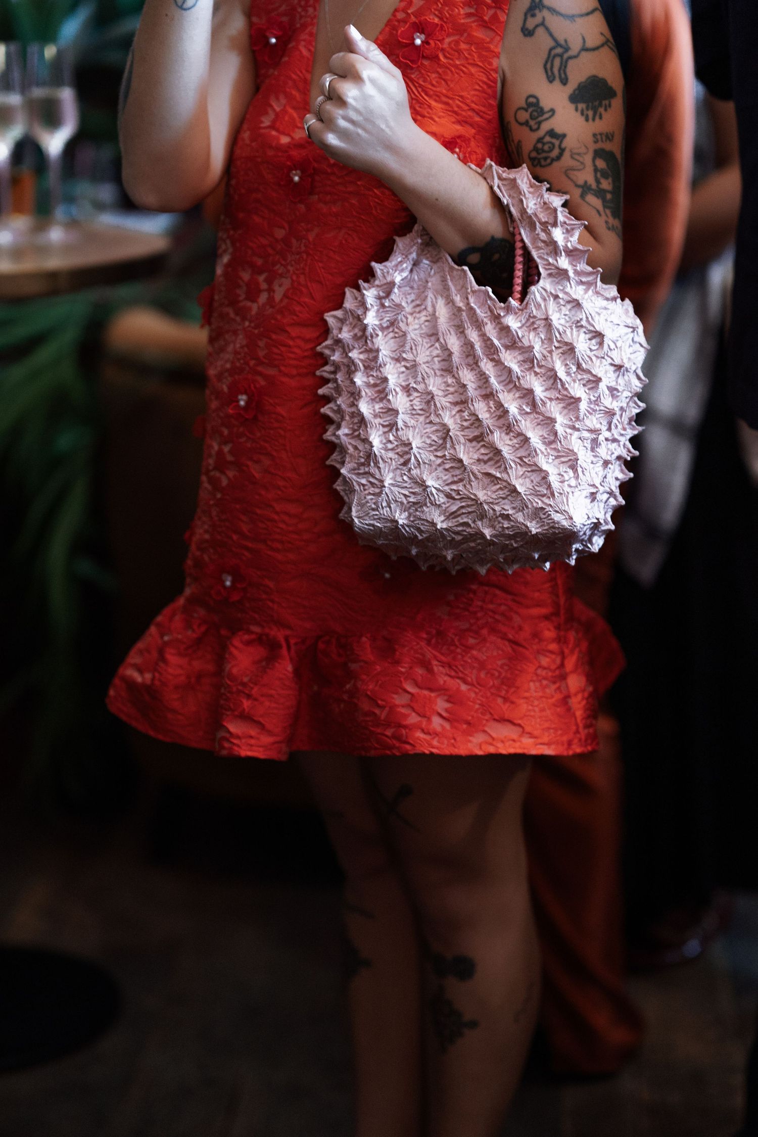 Woman in red lace dress holding a white ruffled handbag, with visible tattoos on arms and legs.