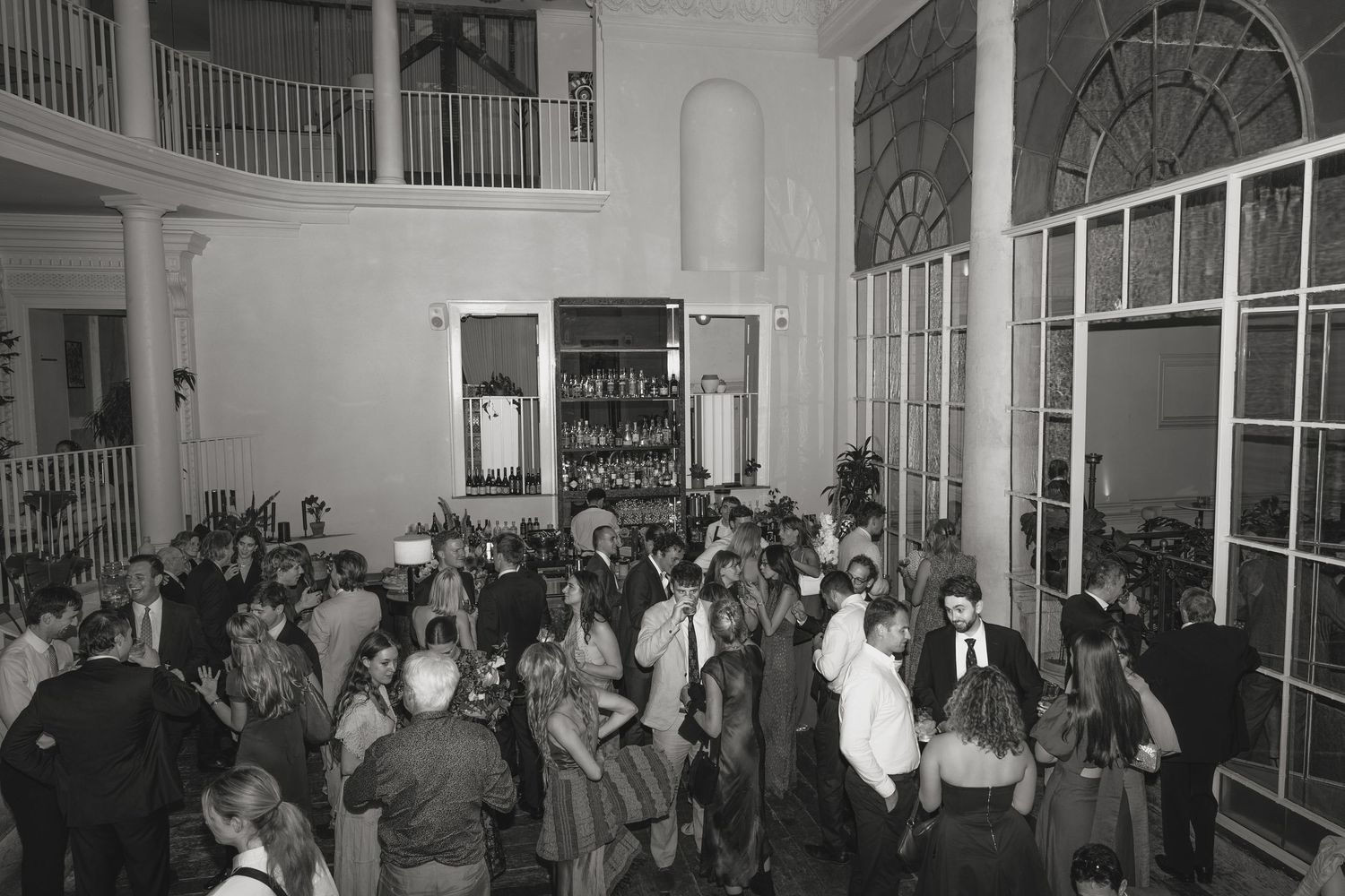 Black and white photo of wedding guests mingling in a grand hall with tall arched windows, a balcony, and a glass-paneled doorway.