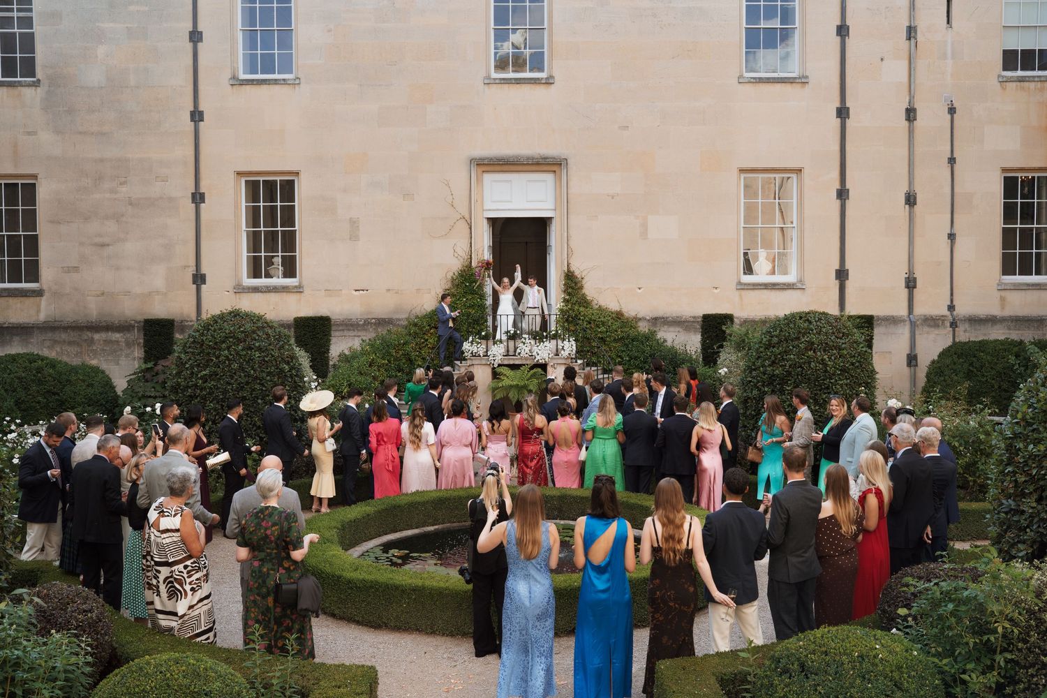 Wedding guests gather outside a grand stone building with manicured gardens for a celebration.