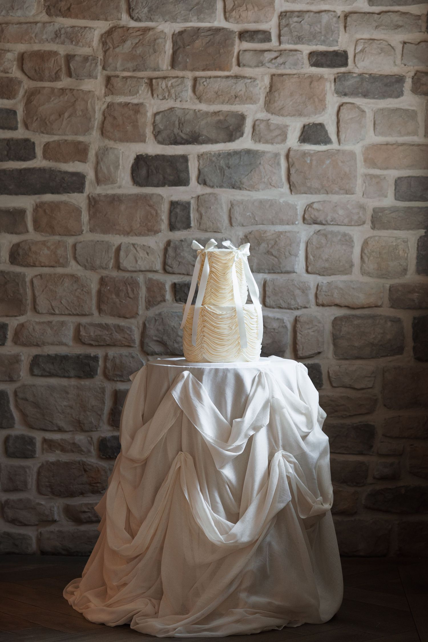 Elegant white wedding cake with draped fabric design displayed on white tablecloth against stone wall backdrop.