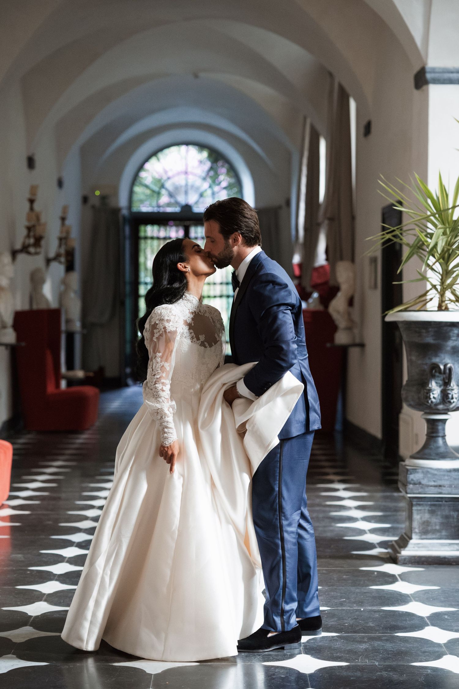 A couple shares an intimate dance in an elegant hallway with arched windows and black and white checkered floors.