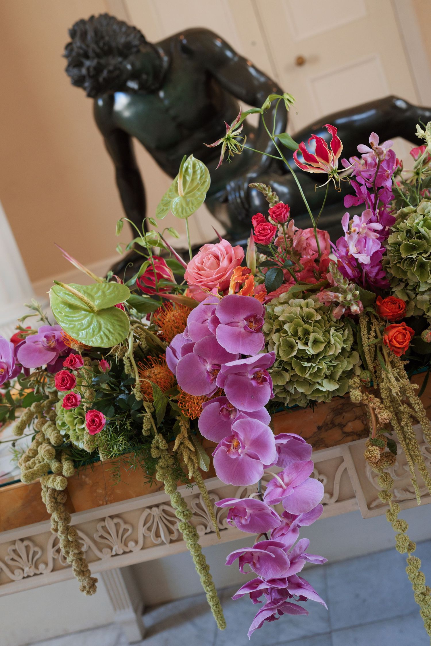 Lush floral arrangement with purple orchids, pink roses, and green hydrangeas displayed before a bronze sculpture.