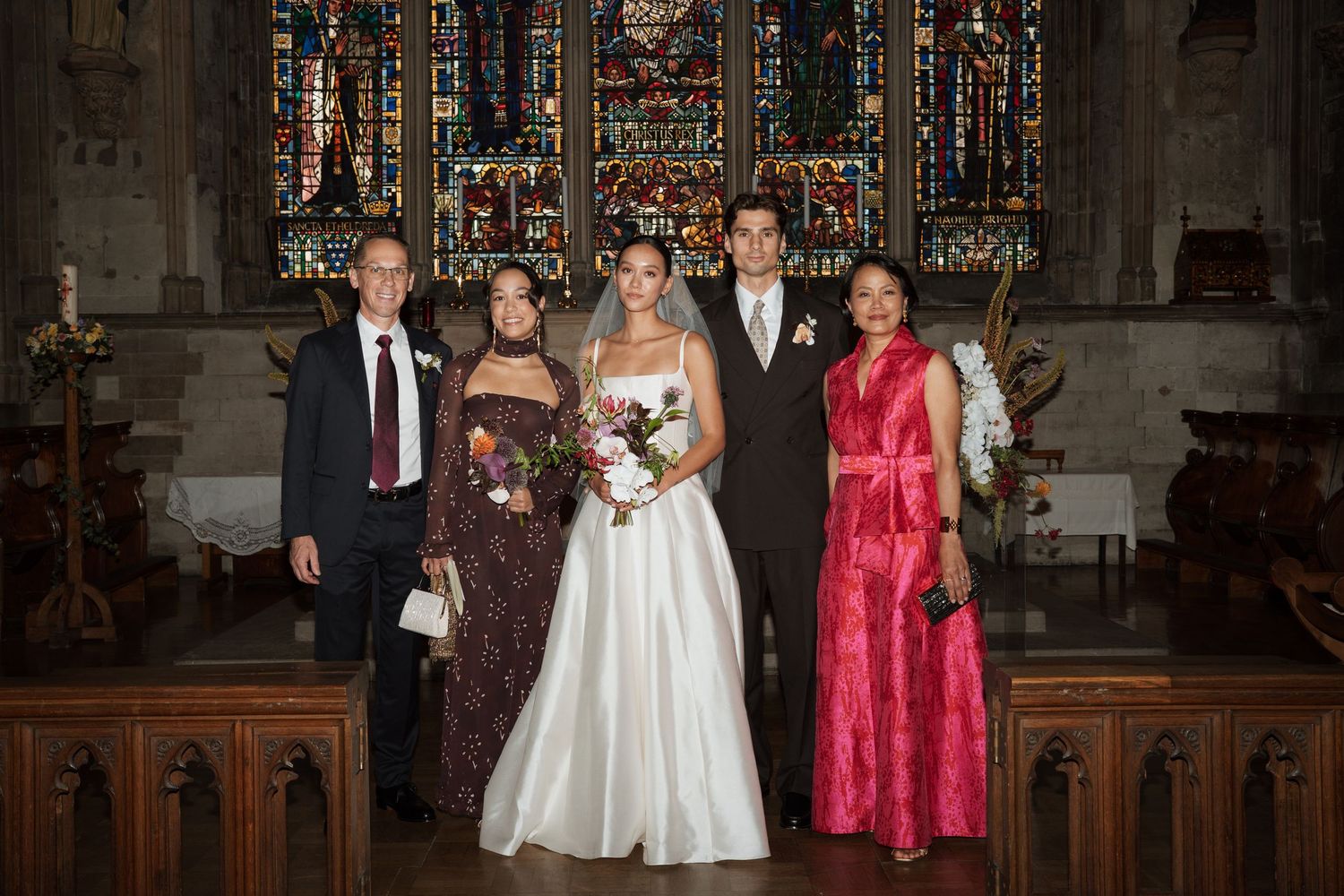 Bride in a white gown holding a bouquet poses with four wedding guests in formal attire inside a church with stained glass windows.