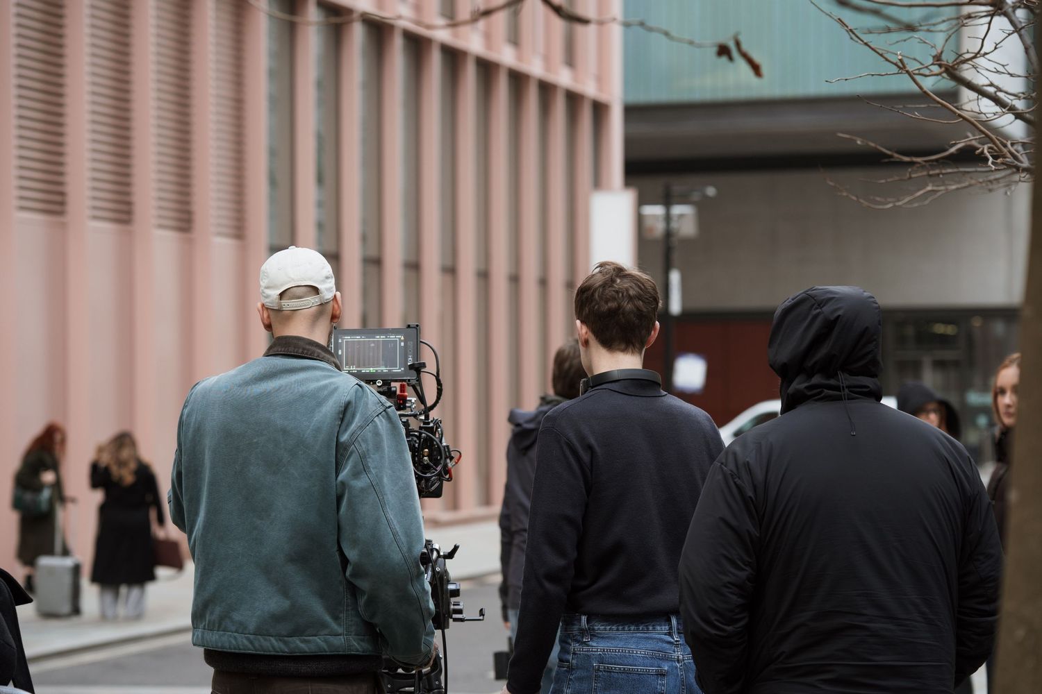 Film crew with camera equipment filming outdoors near a modern pink building on a city street.