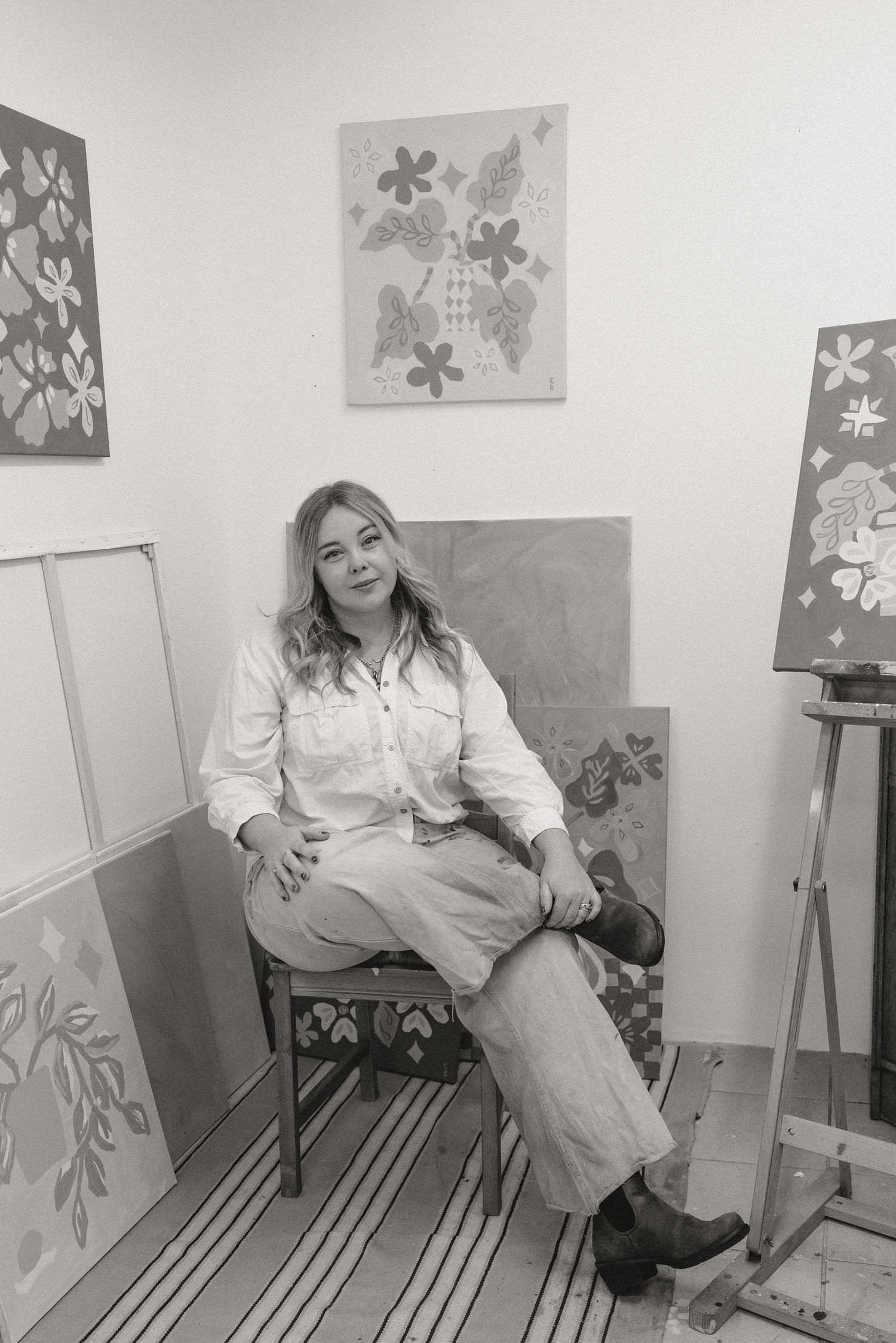 Artist sitting in chair surrounded by floral paintings in black and white studio portrait.