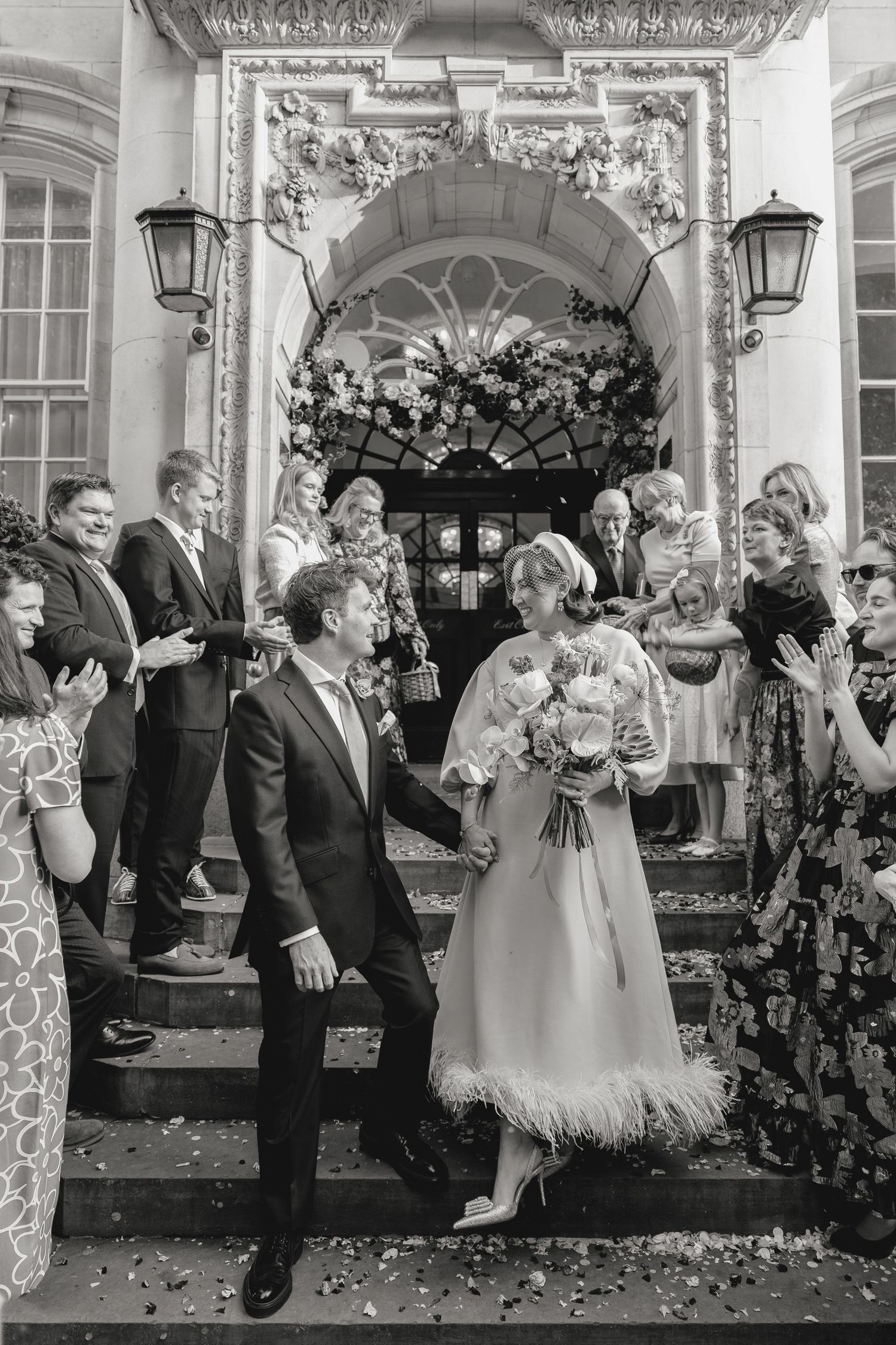 A couple exits a decorated venue while guests throw confetti in this black and white wedding photo.