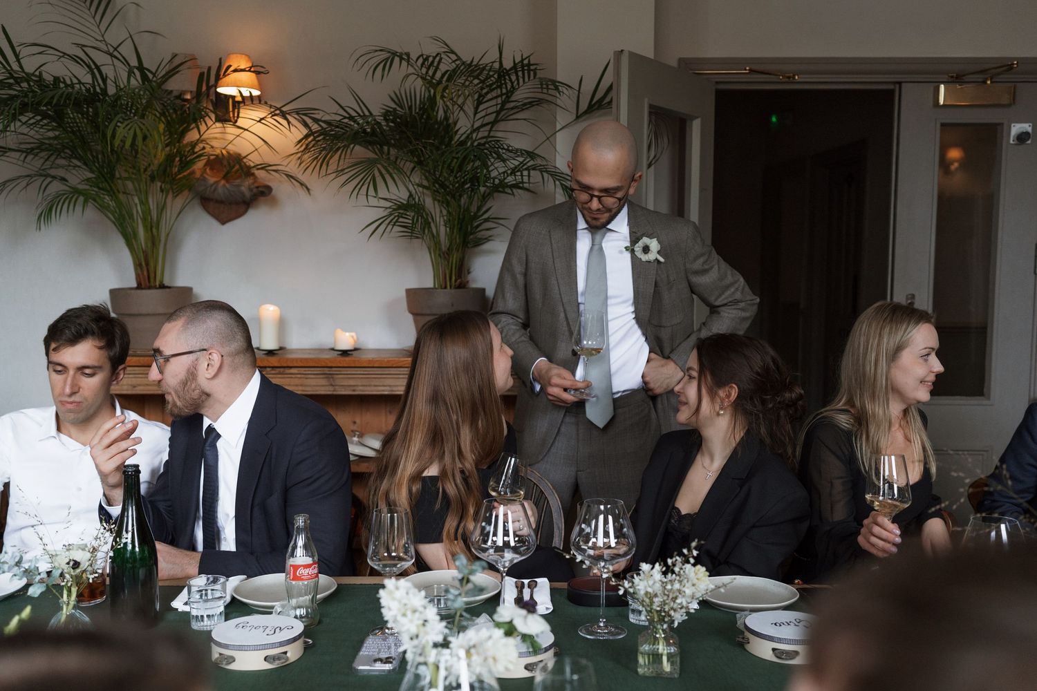 Diners seated at a long table with wine glasses and indoor plants.