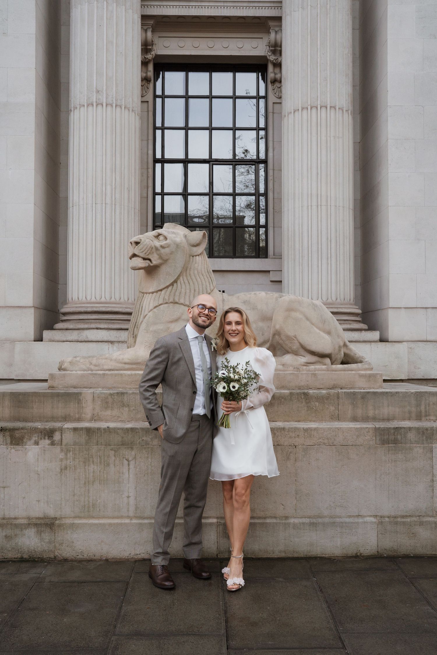Wedding couple posing playfully against neoclassical building with stone lion sculptures.