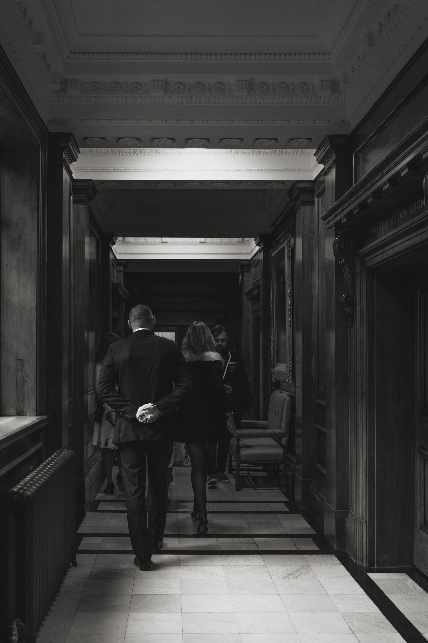A formal couple walks down a dark, elegant hallway with wooden paneling.