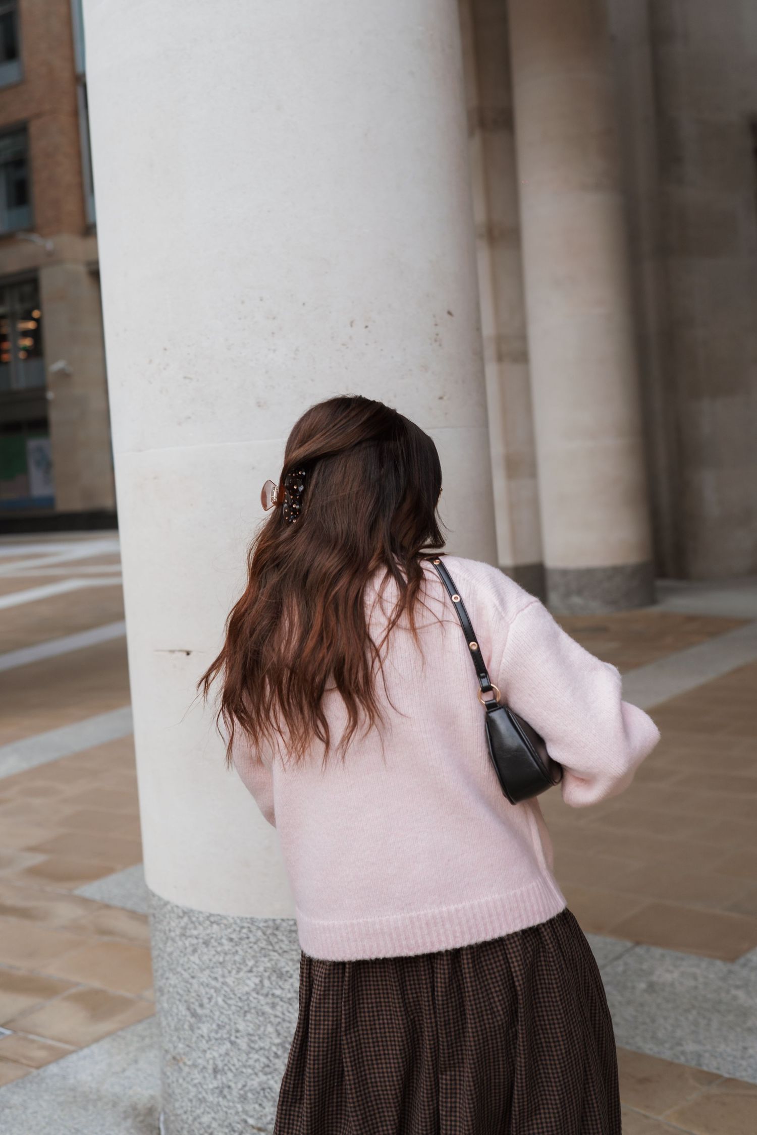 Woman in pink sweater and brown skirt with black bag, standing near large column, viewed from behind.