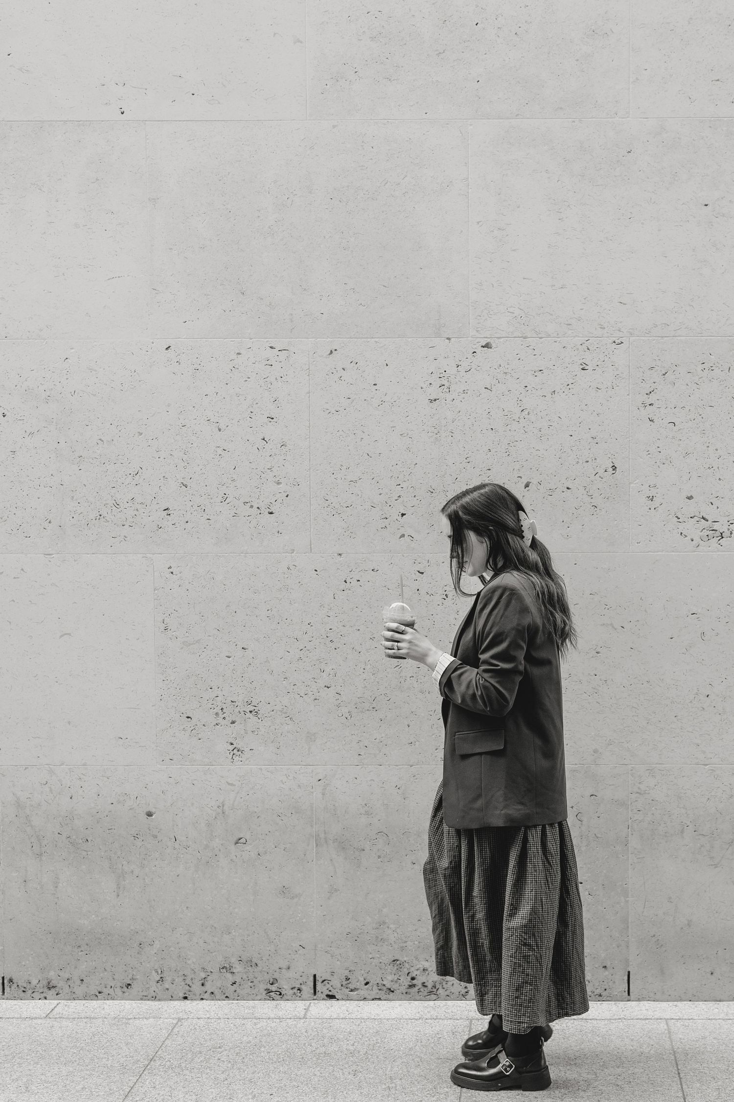 Black and white photo of a woman in a long skirt and blazer holding a drink, standing against a concrete wall.